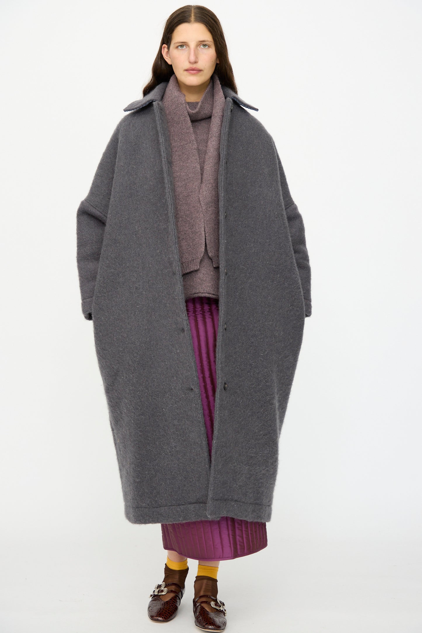 A person stands facing forward in the Cawley Portuguese Hairy Wool Faro Coat in Slate, styled over a mauve top and purple pleated skirt with brown shoes and yellow socks, set against a plain white background.