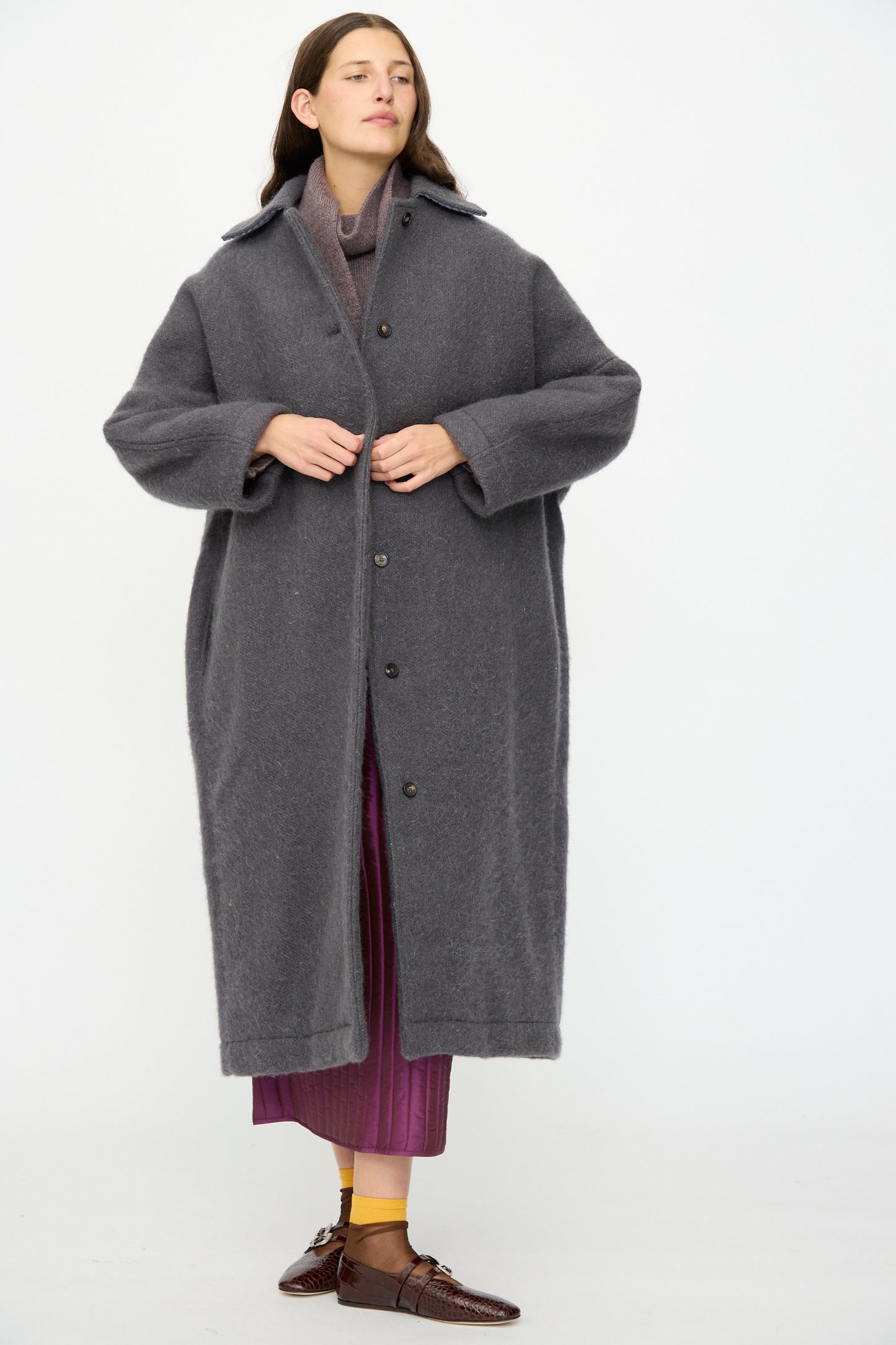 A person wears the Cawley Portuguese Hairy Wool Faro Coat in Slate—an oversized piece made in London—over a purple outfit, standing against a plain white background.