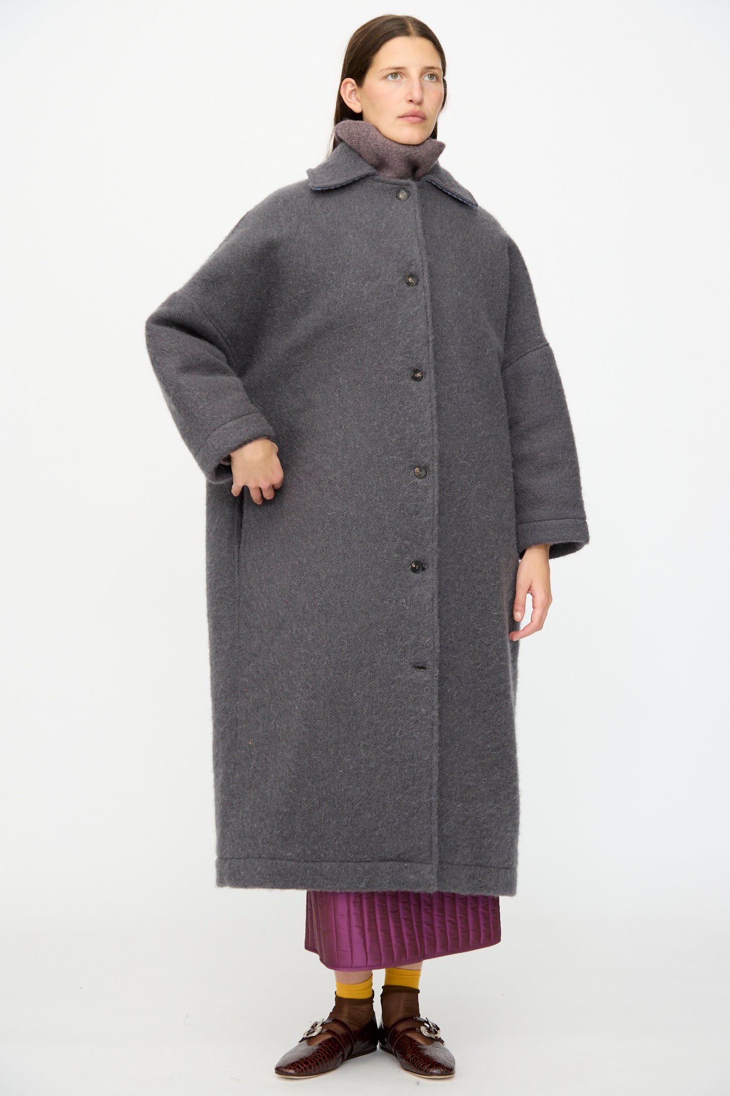 A person stands against a white background wearing the Cawley Portuguese Hairy Wool Faro Coat in Slate, buttoned over a pink skirt, paired with brown shoes and yellow socks.