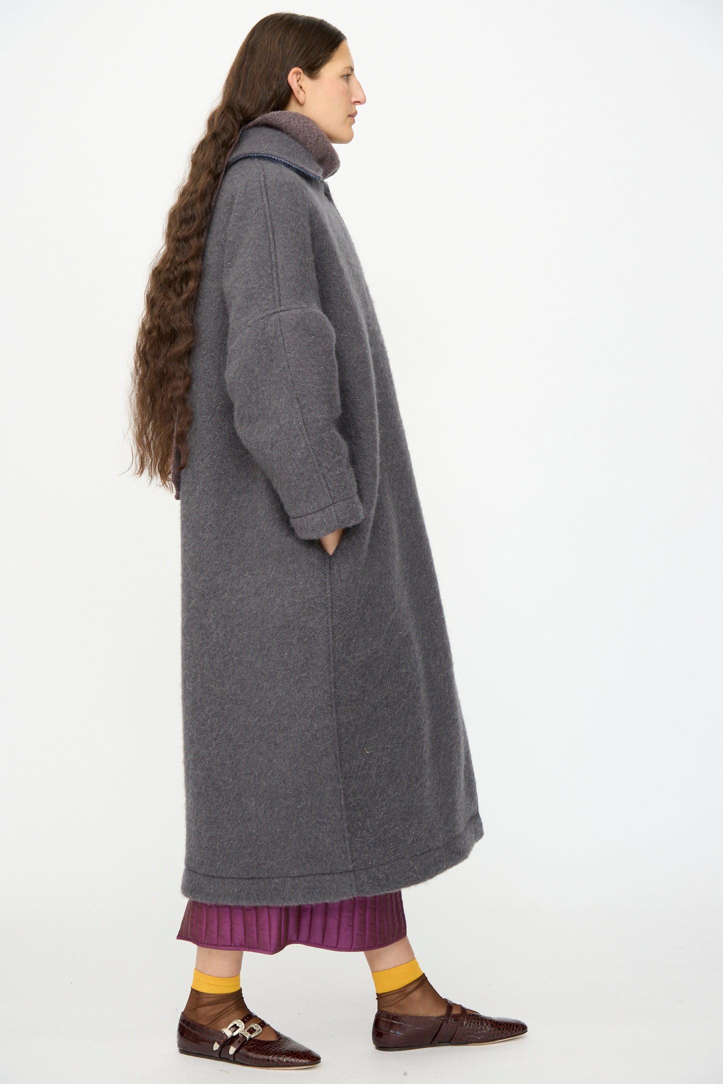 A person with long hair stands sideways in the Cawley Portuguese Hairy Wool Faro Coat in Slate, paired with a purple skirt, yellow socks, and brown shoes against a white background.