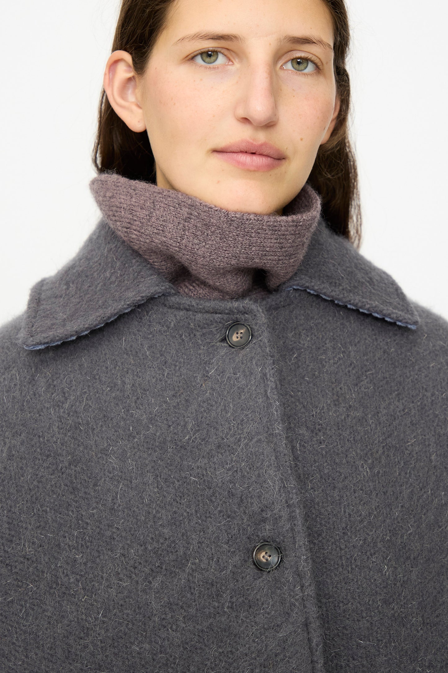 A person with long dark hair wears the Cawley Portuguese Hairy Wool Faro Coat in Slate, an oversized style with large buttons, over a high-necked mauve knit sweater.
