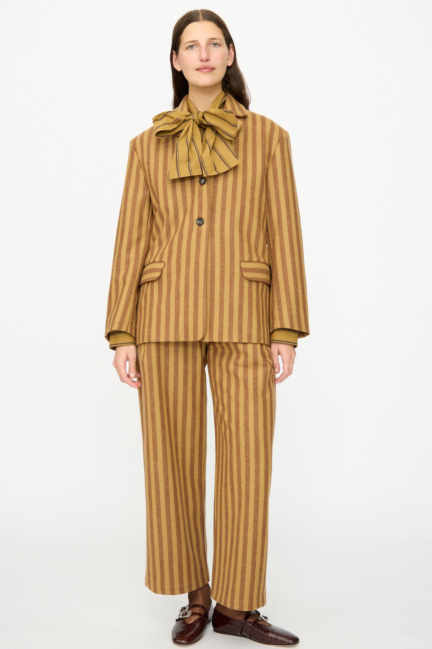 A person stands against a plain background wearing Cawley’s Portuguese Striped Wool Andie Blazer in Goldfinch and Bordeaux, styled with wide legs, a matching neck bow, and brown shoes—an effortlessly chic look made in London.