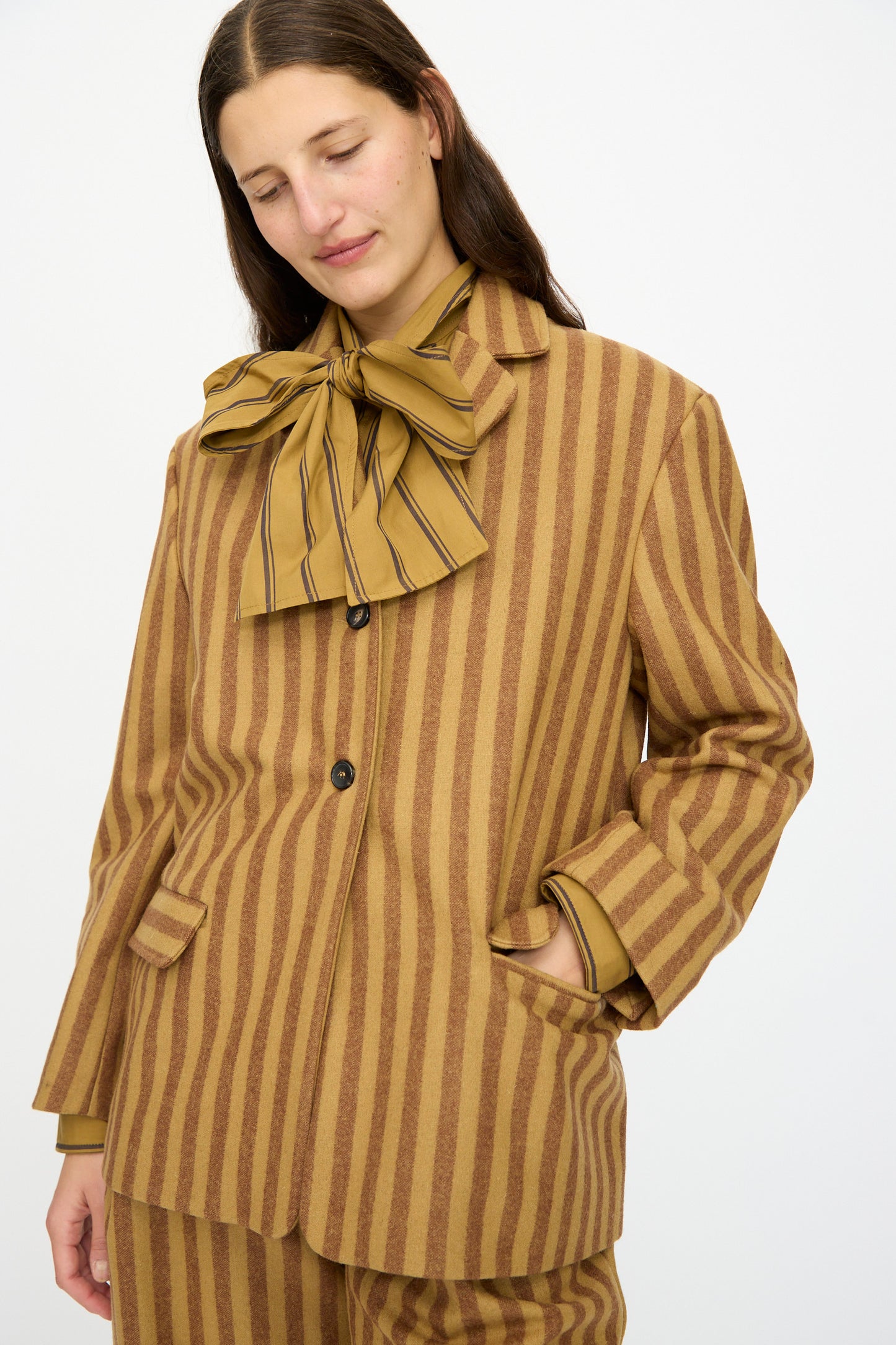 A person wearing the Cawley Portuguese Striped Wool Andie Blazer in Goldfinch and Bordeaux, featuring brown and tan vertical stripes with a large matching bow, stands with one hand in their pocket and looks down. Made in London.