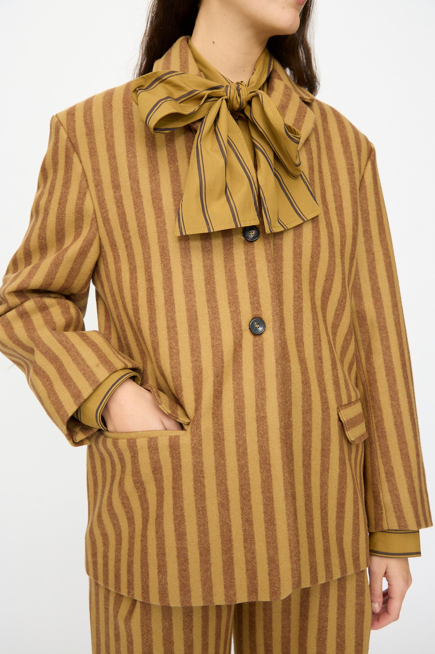 Someone wearing the Cawley Portuguese Striped Wool Andie Blazer in Goldfinch and Bordeaux, featuring brown and tan vertical stripes, stands with one hand in their pocket and a matching bow tie shirt.