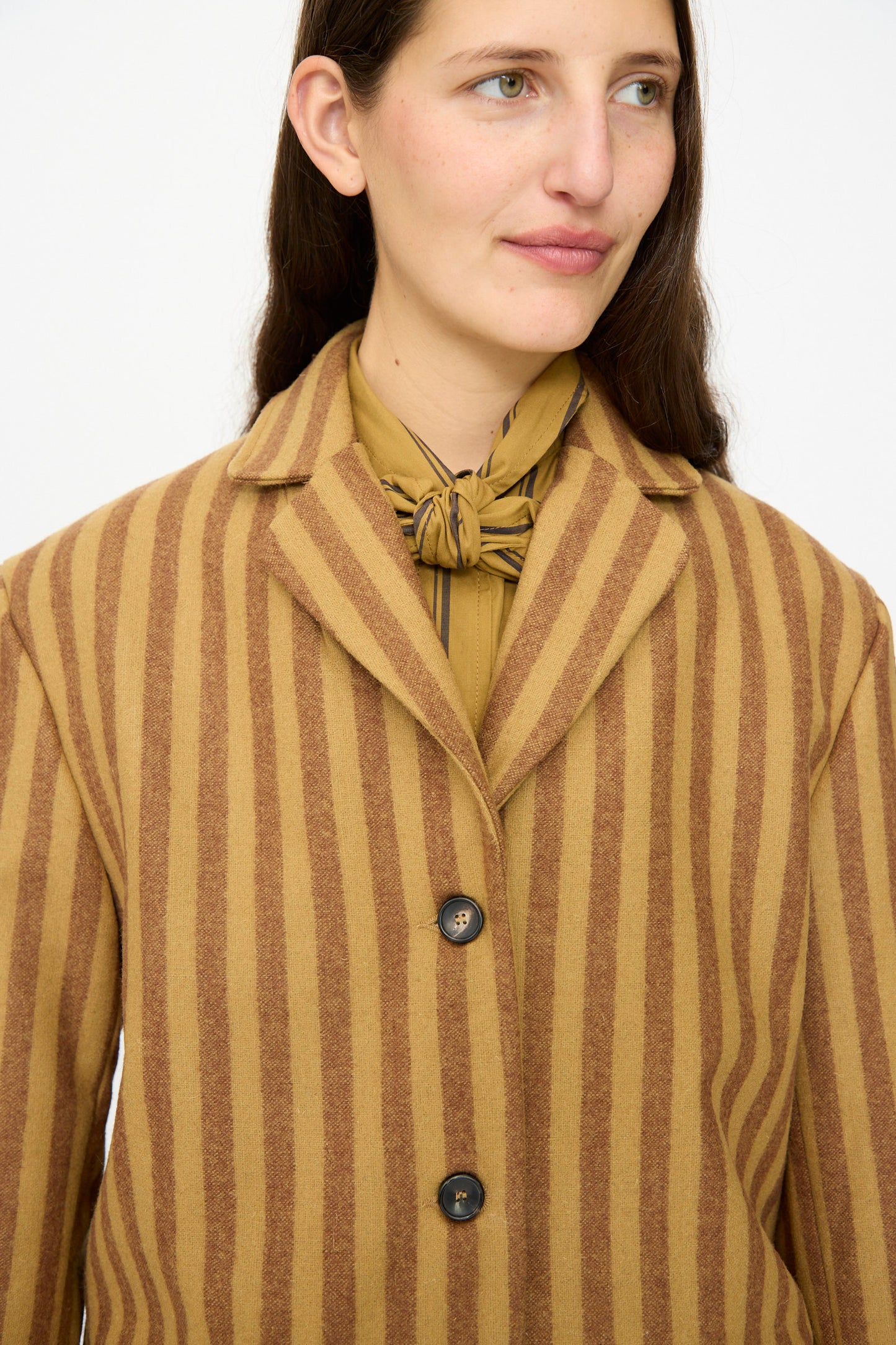 A person wears the Cawley Portuguese Striped Wool Andie Blazer in Goldfinch and Bordeaux over a tan bow-tied blouse, standing against a plain background.