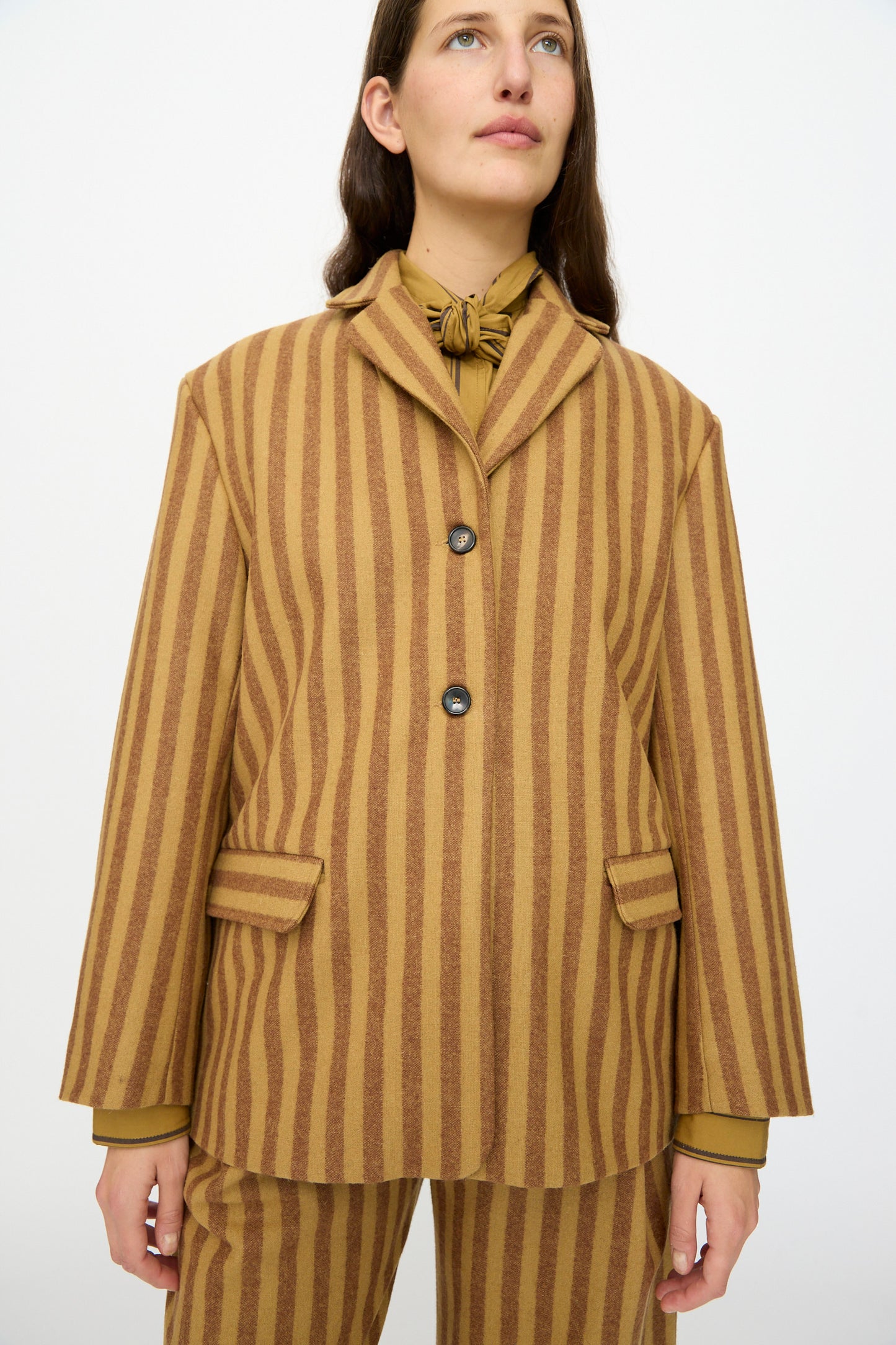 A person wears the Cawley Portuguese Striped Wool Andie Blazer in Goldfinch and Bordeaux with matching pants, a collared shirt, and a necktie while standing against a plain white background.