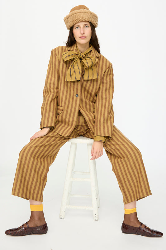 A person sits on a white stool in a Cawley Portuguese Striped Wool Andie Blazer in Goldfinch and Bordeaux, paired with a large bow tie, beige fur hat, yellow socks, and brown shoes against a plain background. The blazer is made in London.