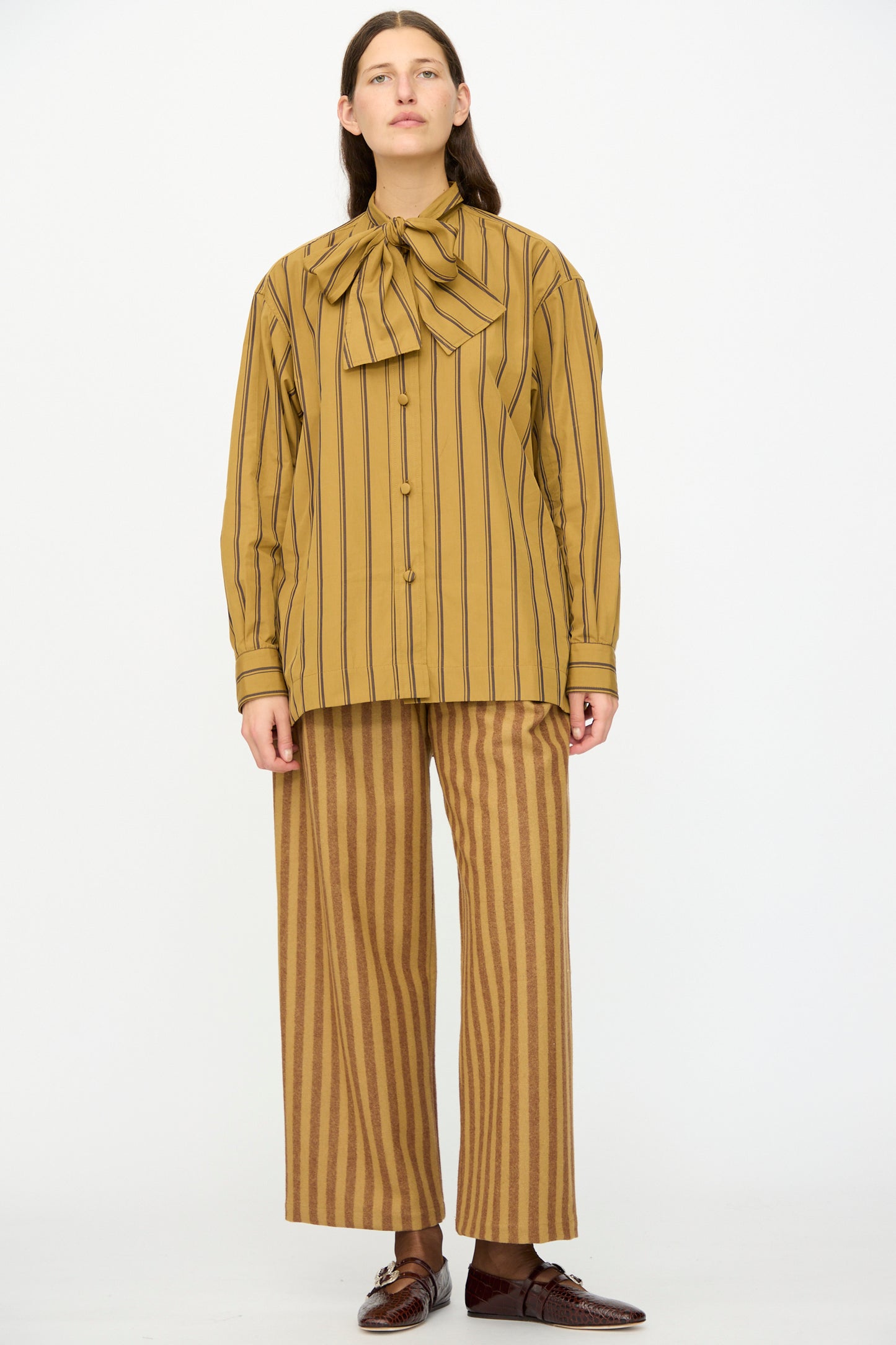 A person stands against a white background wearing Cawley's Portuguese Striped Wool Georgia Trouser in Goldfinch and Bordeaux, paired with a mustard yellow vertically striped blouse with a large bow and brown shoes.