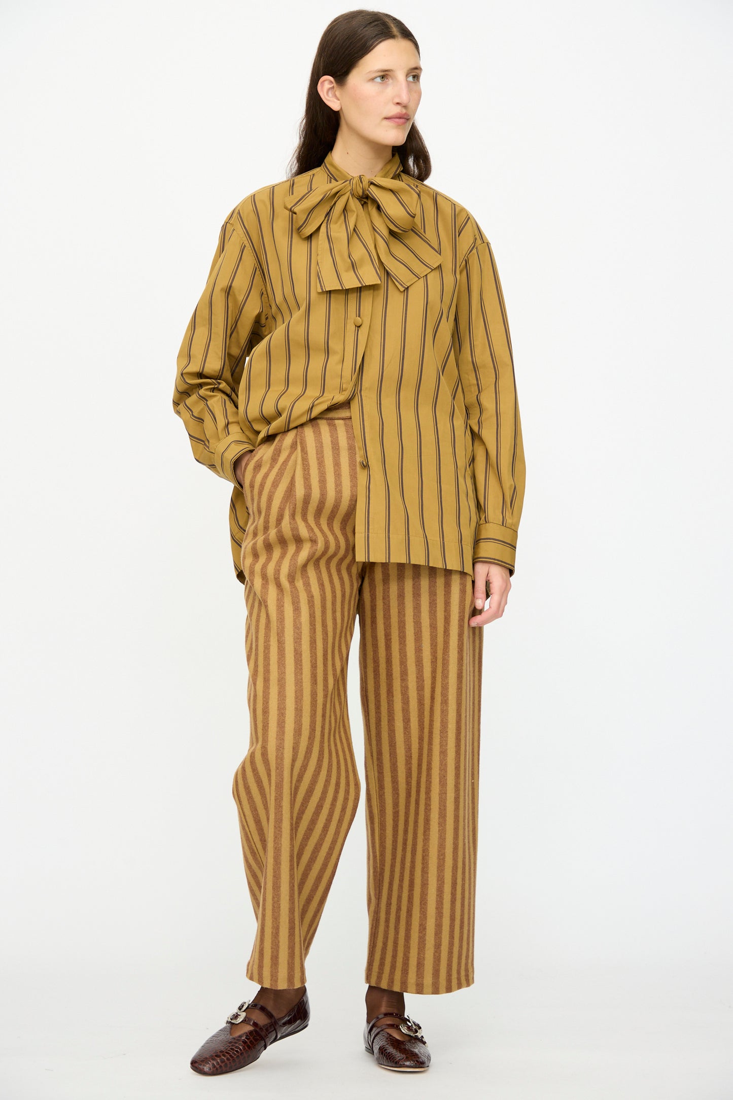 A woman wears Cawley's Portuguese Striped Wool Georgia Trouser in Goldfinch and Bordeaux, paired with a loose mustard striped blouse featuring a large neck bow and brown shoes.