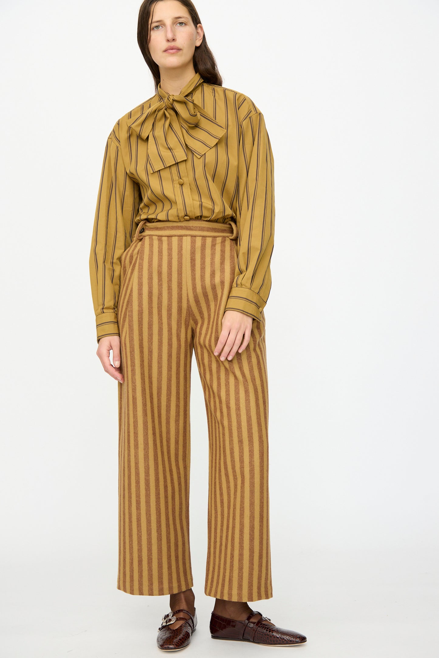 A person stands against a white background wearing Cawley's Portuguese Striped Wool Georgia Trouser in Goldfinch and Bordeaux, paired with a mustard striped blouse and brown loafers.
