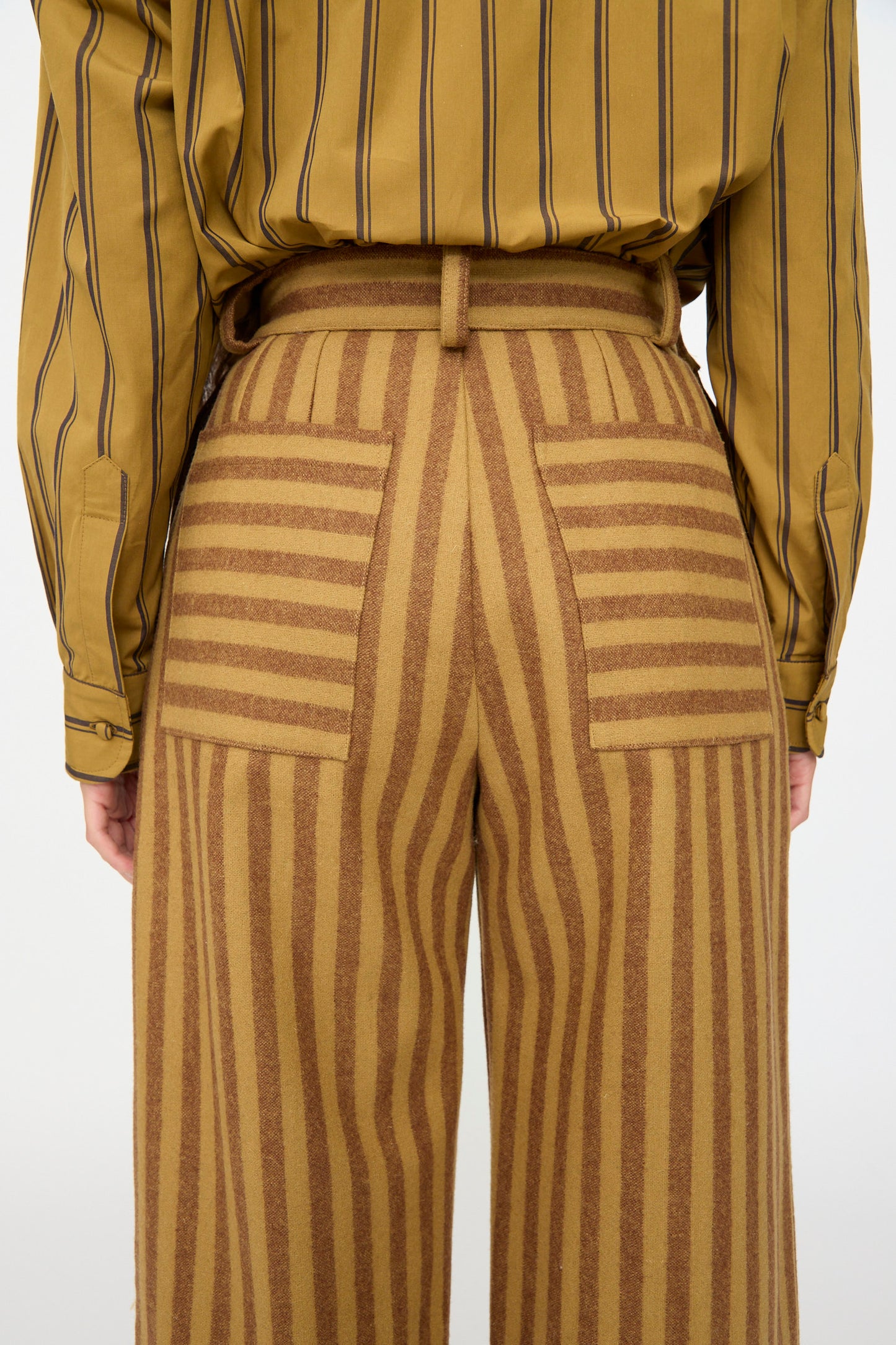 A person viewed from the back wears Cawley's Portuguese Striped Wool Georgia Trouser in Goldfinch and Bordeaux, featuring high waist and horizontal and vertical stripes, paired with a mustard yellow striped shirt. Made in London.