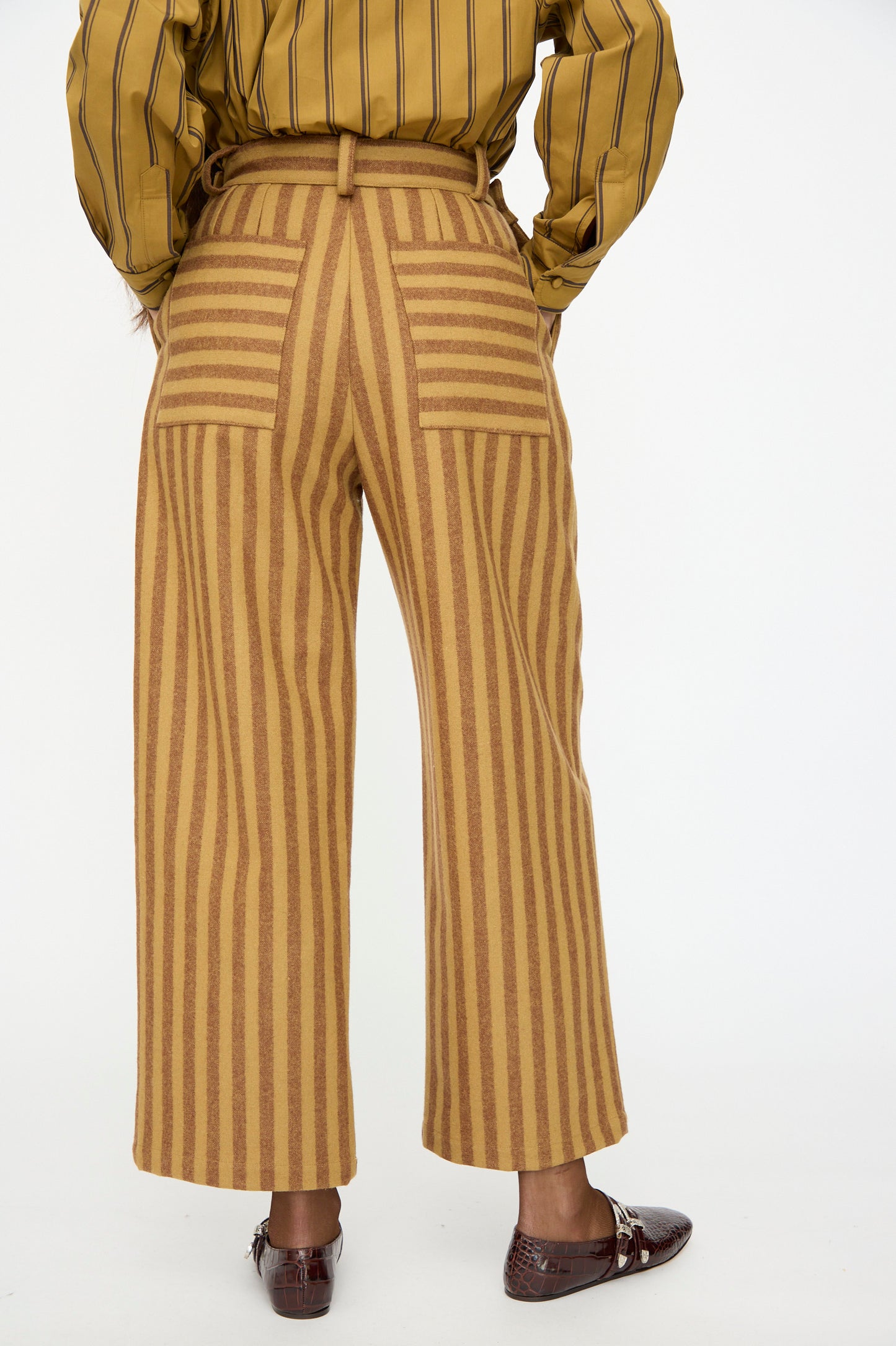 Person wearing Cawley's Portuguese Striped Wool Georgia Trouser in Goldfinch and Bordeaux, paired with a mustard yellow striped shirt and brown loafers, stands with back to camera; a stylish London-made look.