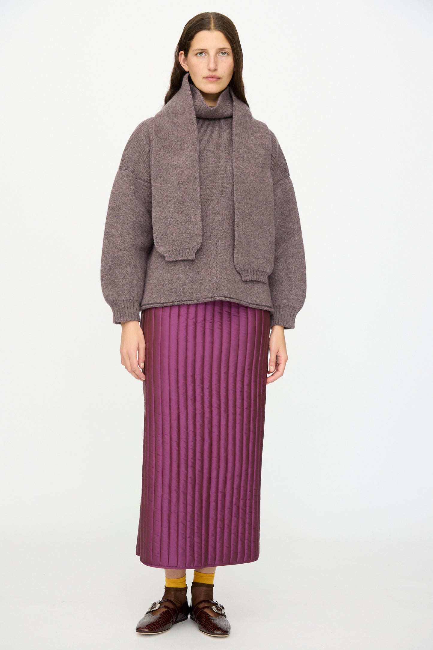 A woman faces forward wearing a mauve sweater with a matching scarf, the Cawley Quilted Silk Sadie Skirt in Plum, yellow socks, and brown shoes against a plain white background.