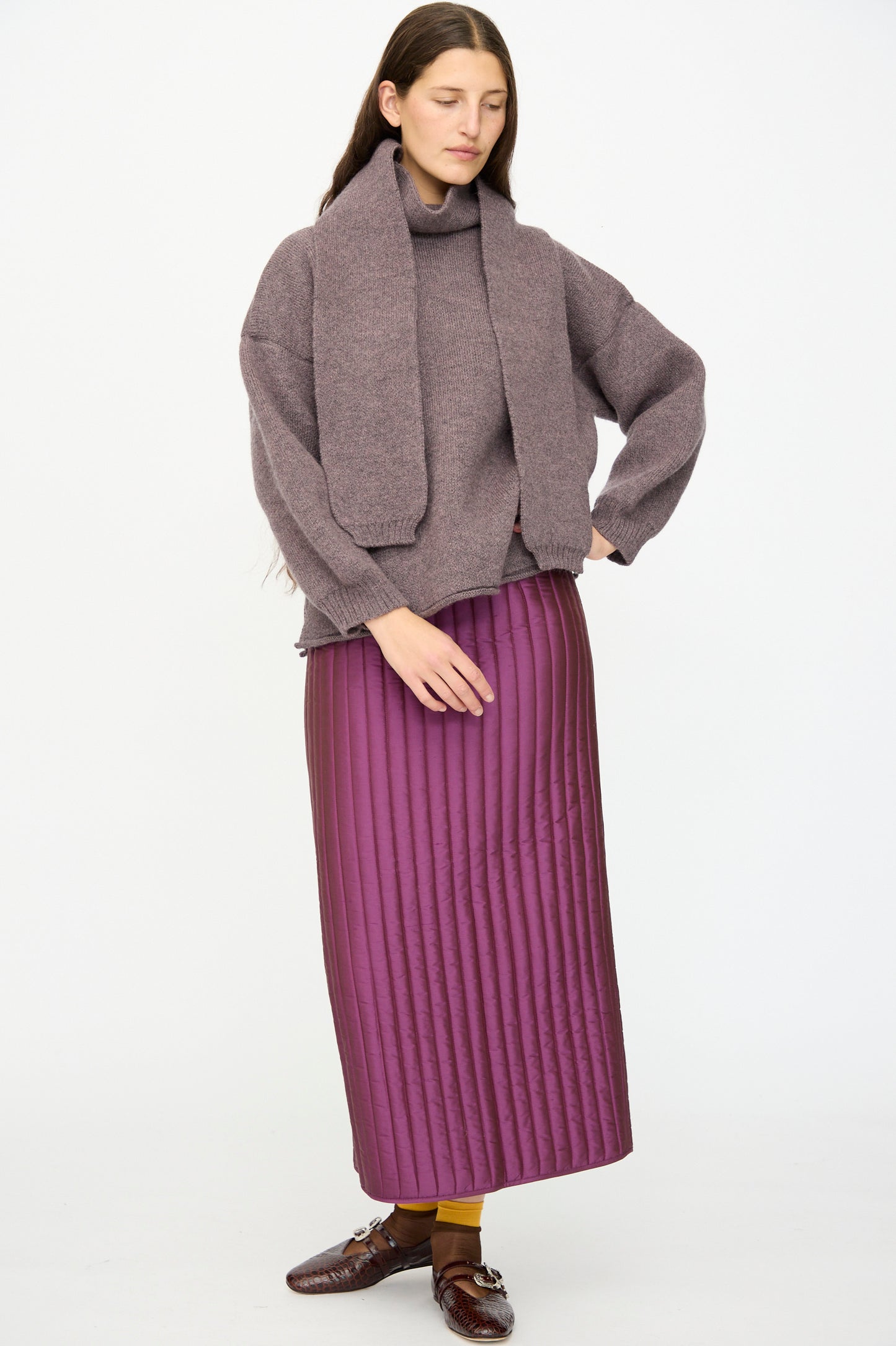 A woman stands against a plain background wearing a purple knit sweater with matching scarf, brown shoes, mustard-yellow socks, and the Cawley Quilted Silk Sadie Skirt in Plum.
