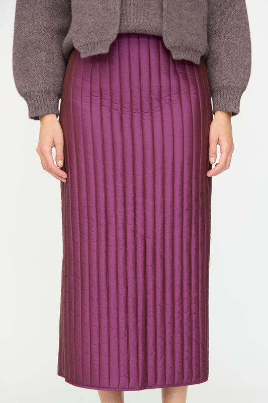 A person wears the Cawley Quilted Silk Sadie Skirt in Plum, a high-waisted pencil skirt made in London with vertical stitching, paired with a brown sweater, standing against a plain background.