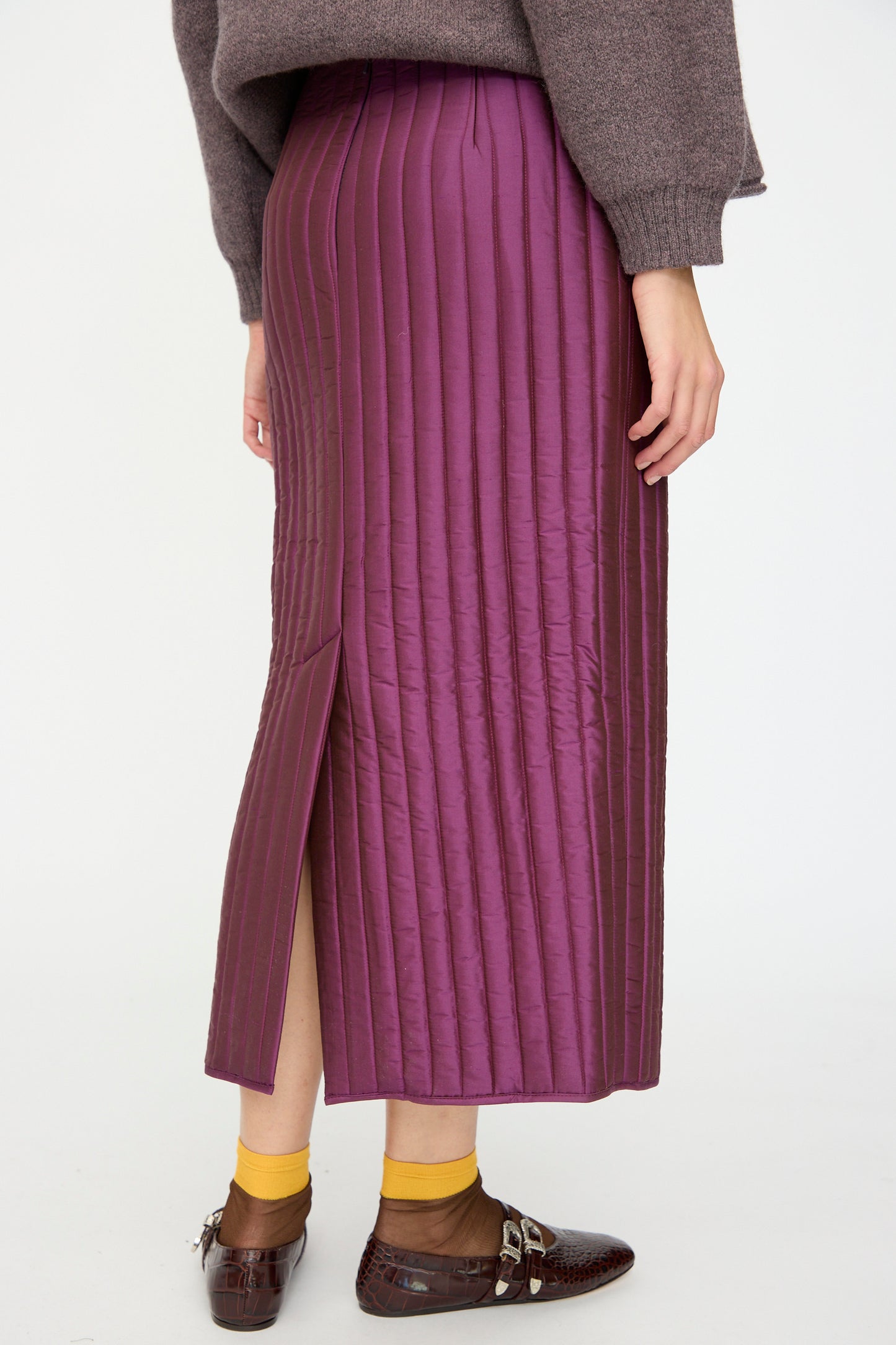 A person wearing the Cawley Quilted Silk Sadie Skirt in Plum, a brown sweater, yellow socks, and brown patent shoes.