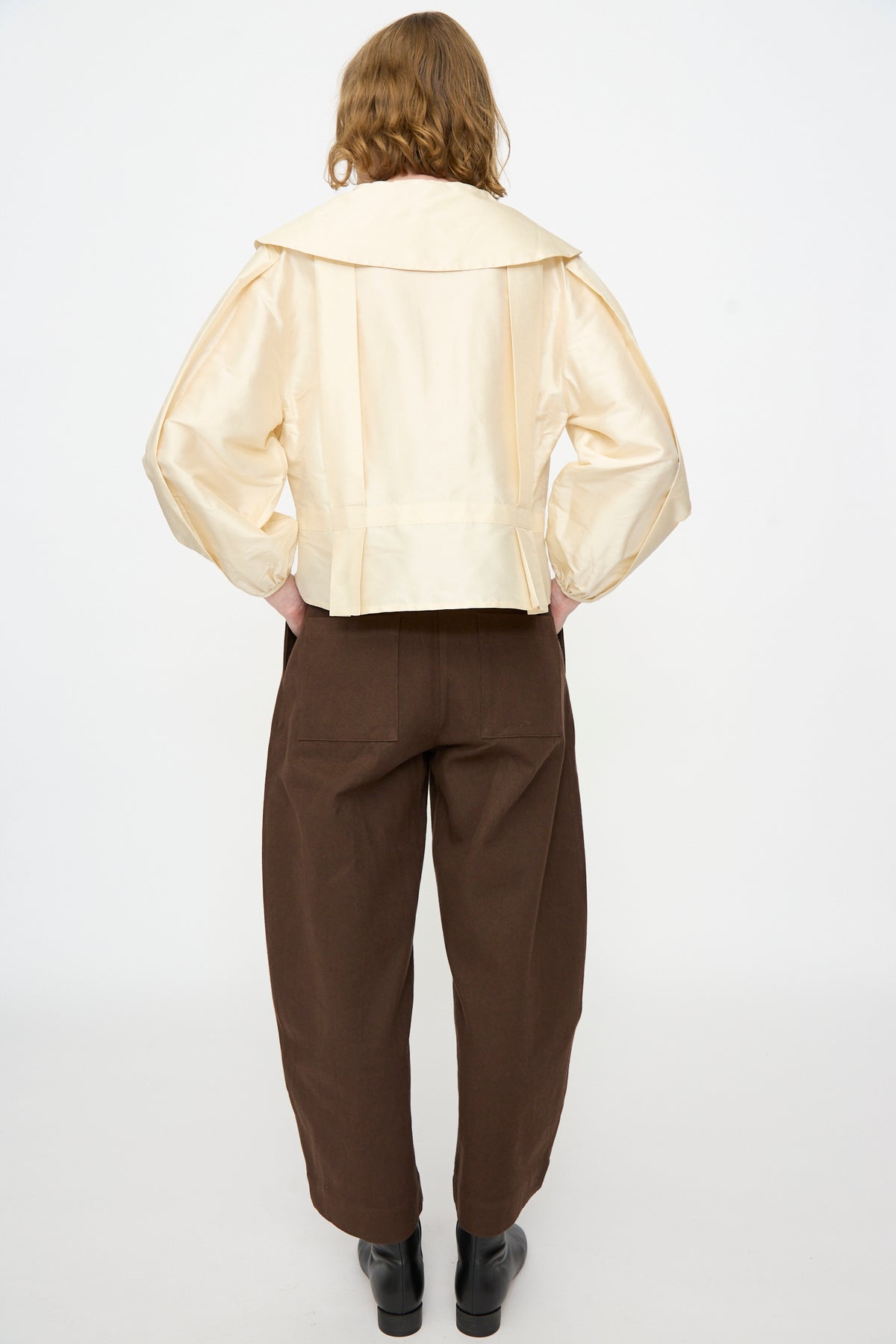 A person stands with their back to the camera, dressed in a cream-colored jacket featuring a wide collar and loose brown pants against a plain white backdrop. The ensemble is elevated by the addition of Cawley’s Silk Dupion Box Pleat Blouse in Ivory, subtly layered underneath to lend understated elegance to the look.
