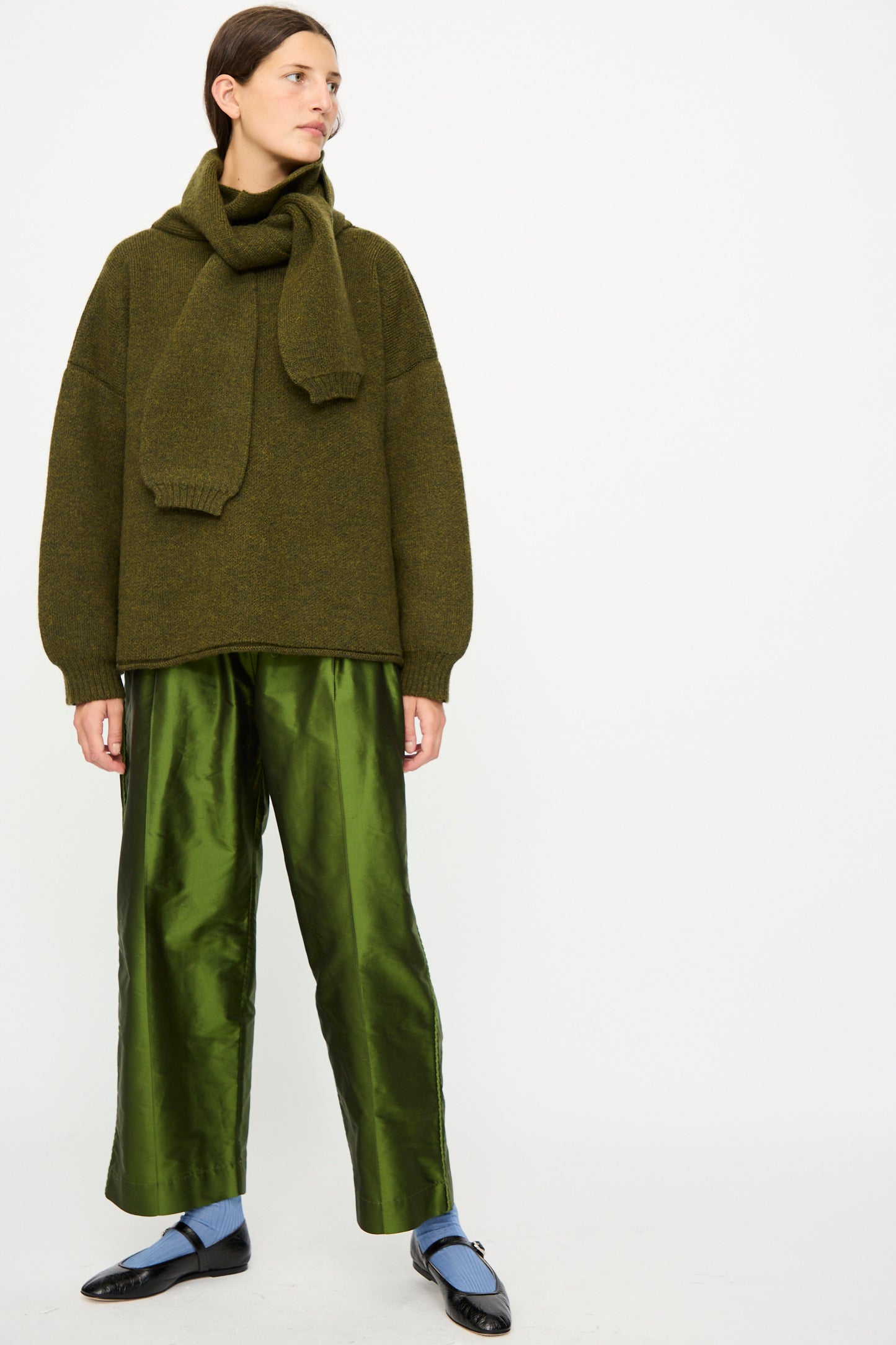 A person wearing the Cawley Silk Dupion Luna Trouser with Trim in Bottle, a green sweater and matching scarf, light blue socks, and black shoes stands against a plain white background.