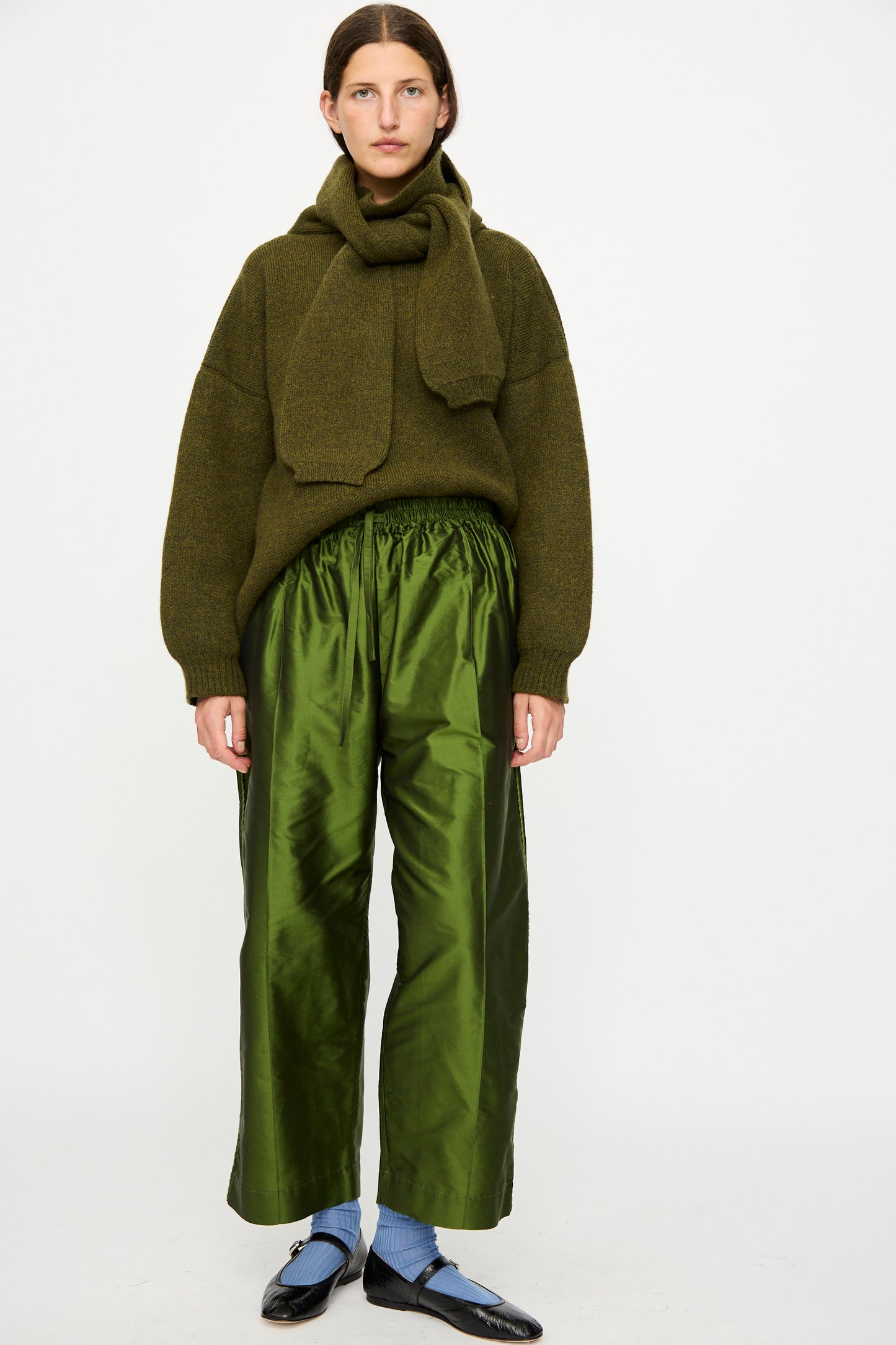 A person stands against a white background, wearing an olive green sweater and matching scarf, blue socks, black shoes, and Cawley’s Silk Dupion Luna Trouser with Trim in Bottle—an ensemble made in London.
