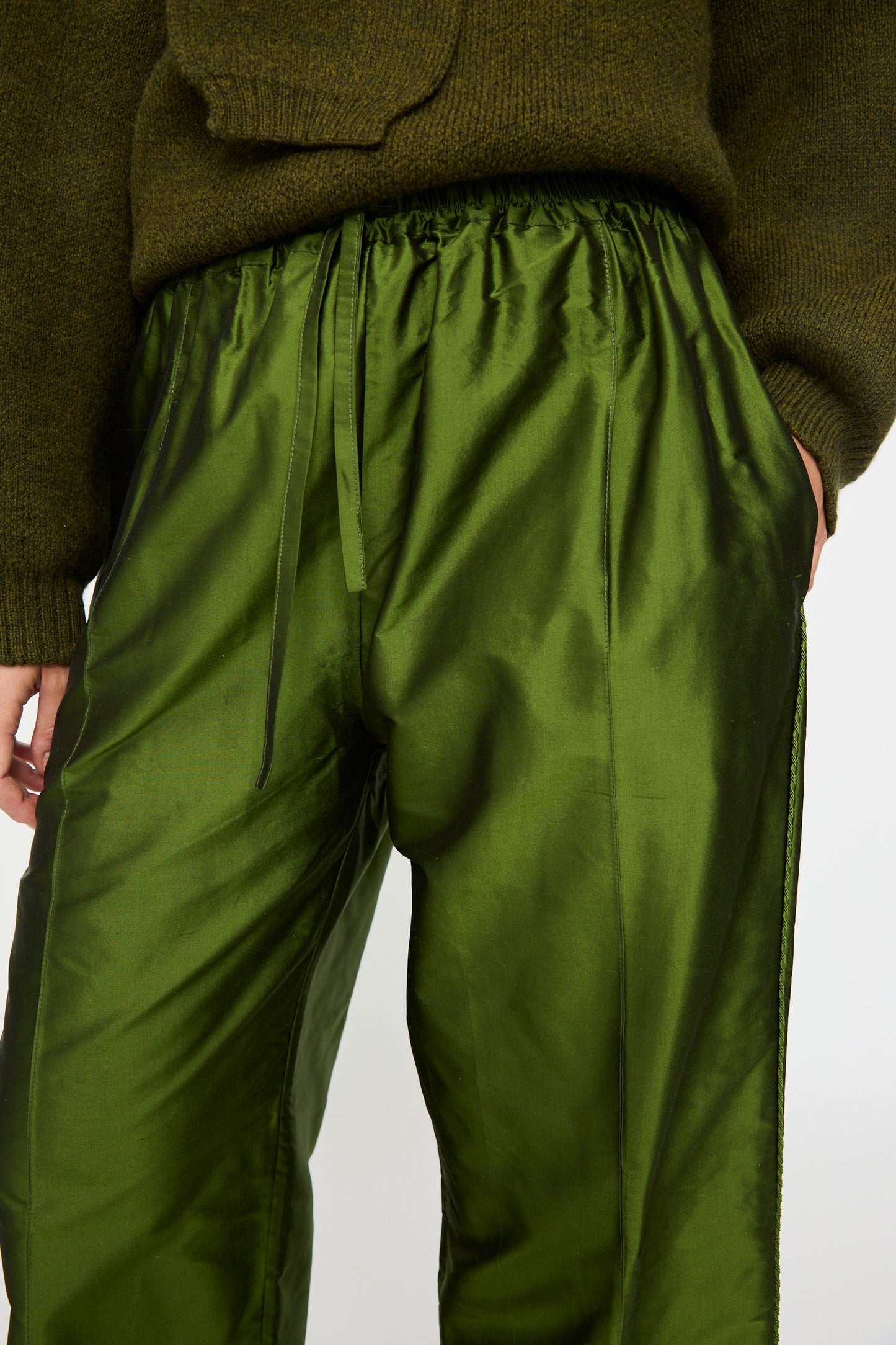 Person wearing Cawley’s Silk Dupion Luna Trouser with Trim in Bottle, paired with a dark green knitted sweater, hands in pockets, shown from midsection to upper thighs.