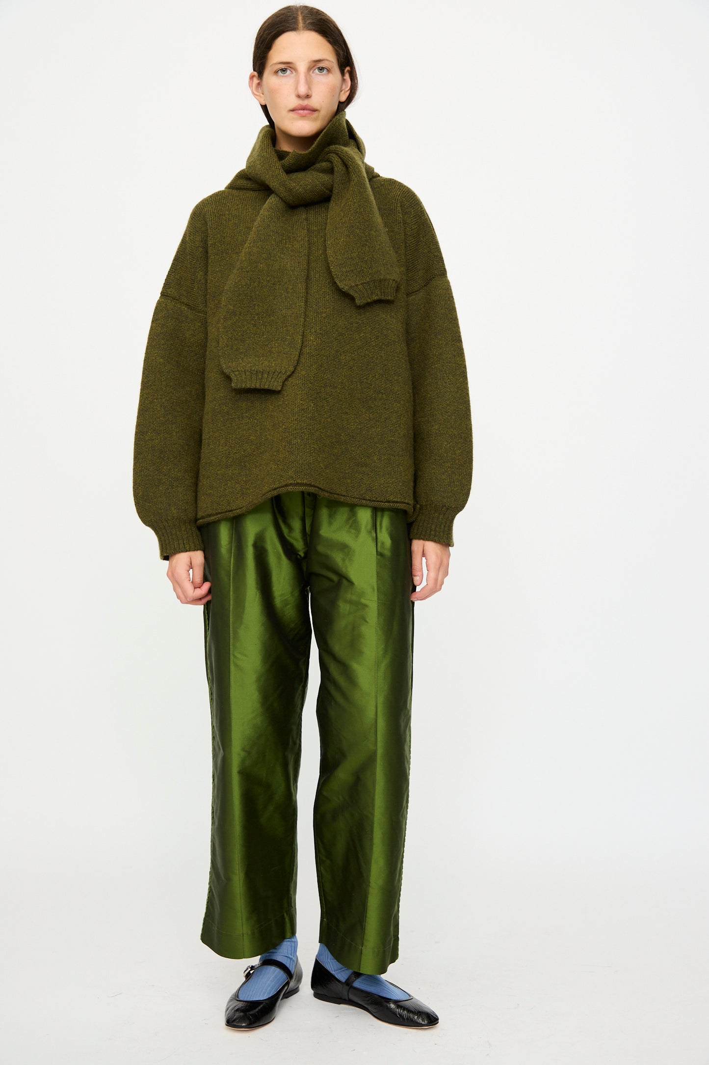 A person stands against a plain background wearing an olive green sweater with a matching scarf, Cawley Silk Dupion Luna Trouser with Trim in Bottle, blue socks, and black shoes.