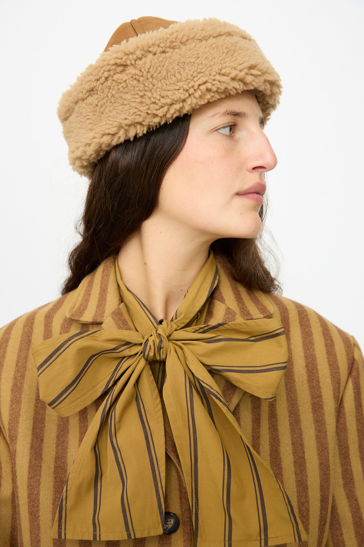 A woman wears the Cawley Suede Back Curly Sheepskin Cap in Tan with a tan and brown striped coat featuring a large bow at the neck, looking to the side against a plain background.