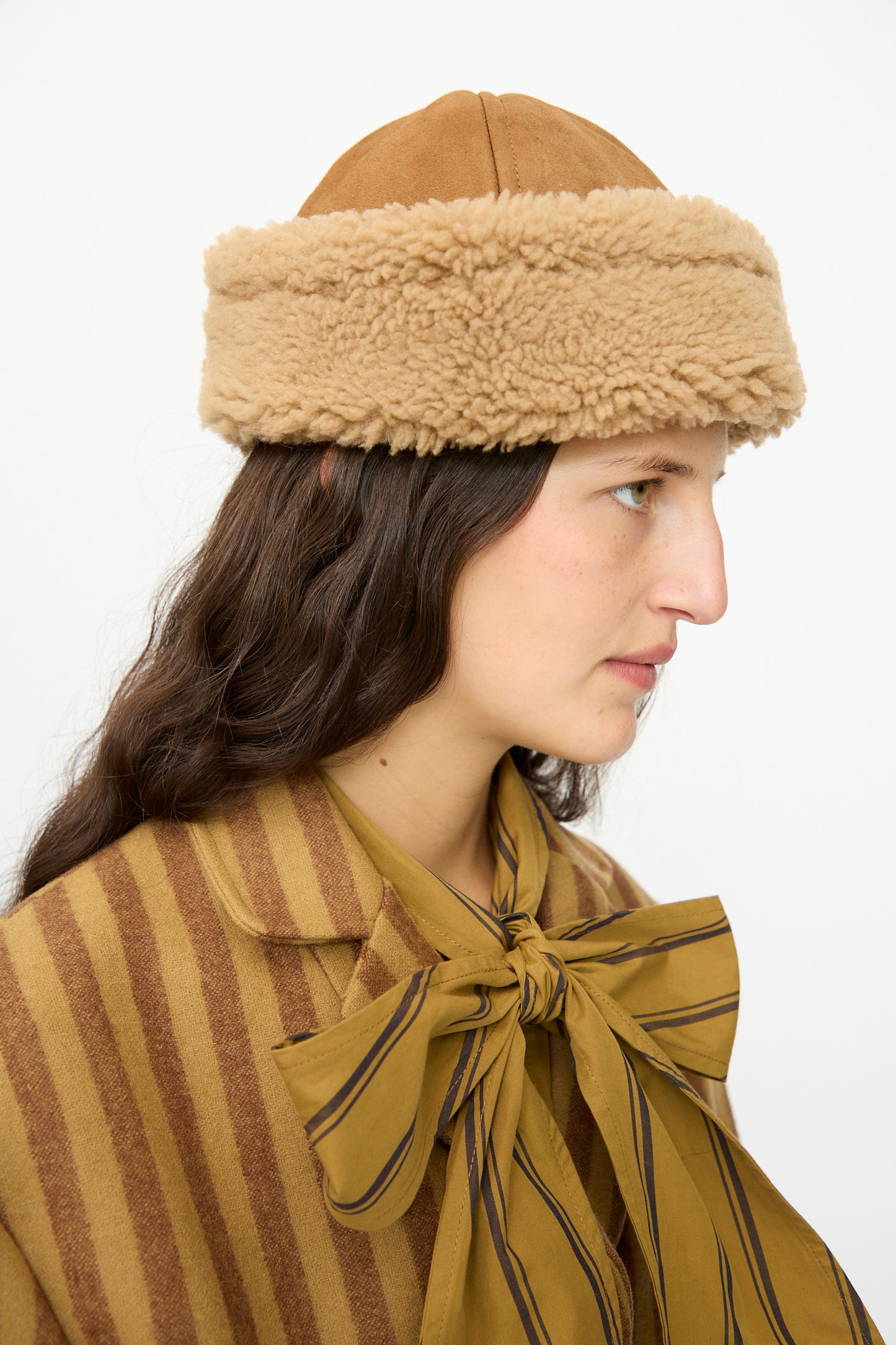 Woman in profile wearing the Cawley Suede Back Curly Sheepskin Cap in Tan and a striped brown coat with a large bow at the collar.