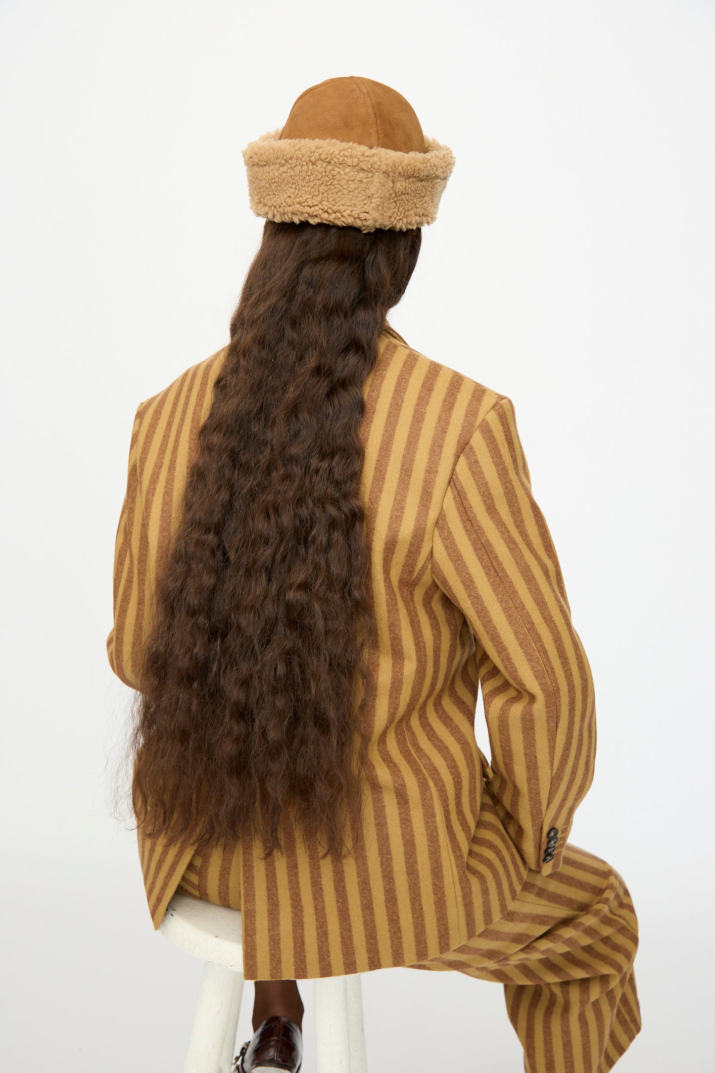 A person with long, wavy brown hair sits on a white stool, wearing a tan and brown striped suit and the Cawley Suede Back Curly Sheepskin Cap in Tan, facing away from the camera.