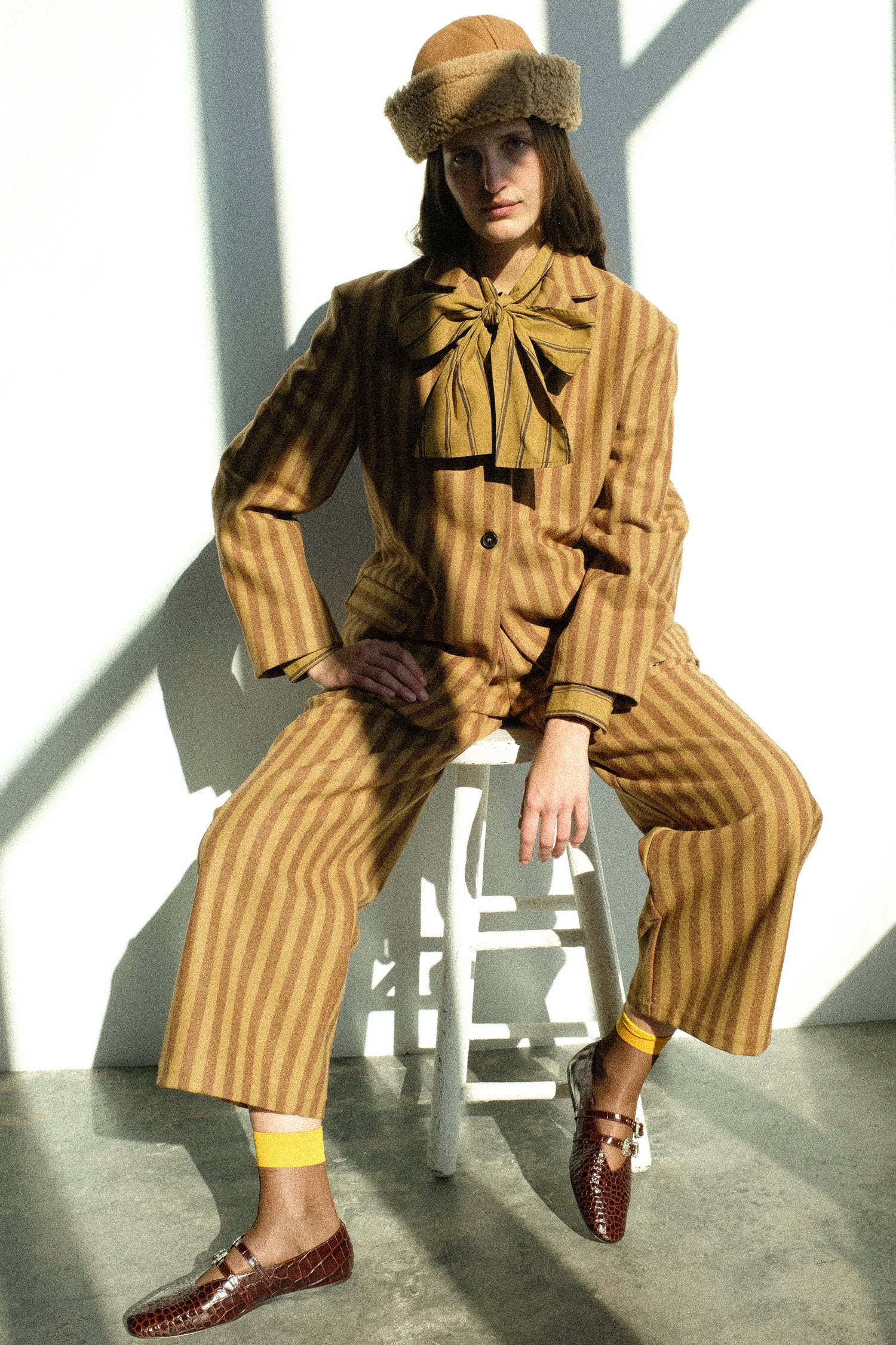 A woman wearing pieces from Cawley's FW25 collection, sitting on a stool against a white background.