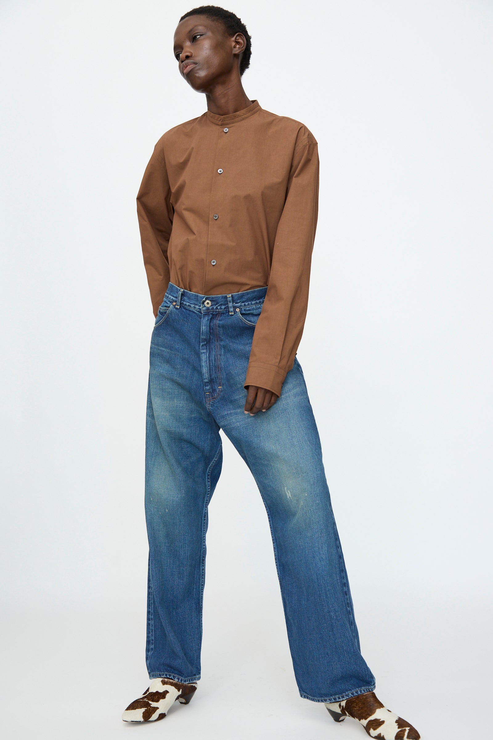 Person standing against a plain background wearing a brown button-up shirt, Chimala 13oz. Selvedge Denim Ranch Pants in Dark Wash, and cow-print shoes.