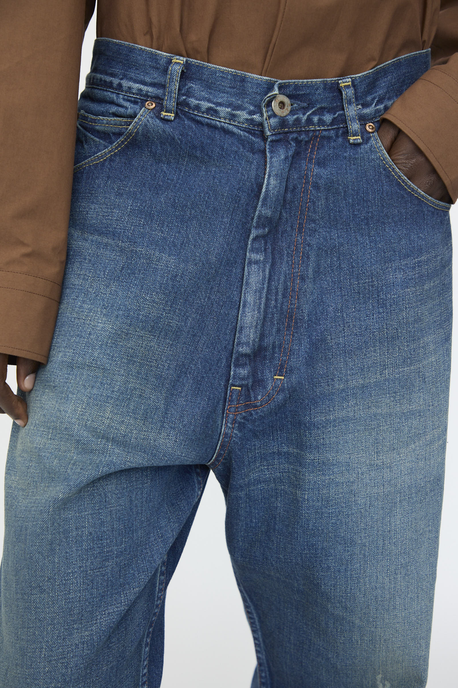 A person wears Chimala 13oz. Selvedge Denim Ranch Pants in Dark Wash—wide-leg, loose-fitting jeans with prominent seams—paired with a brown long-sleeve shirt, one hand in the pocket.