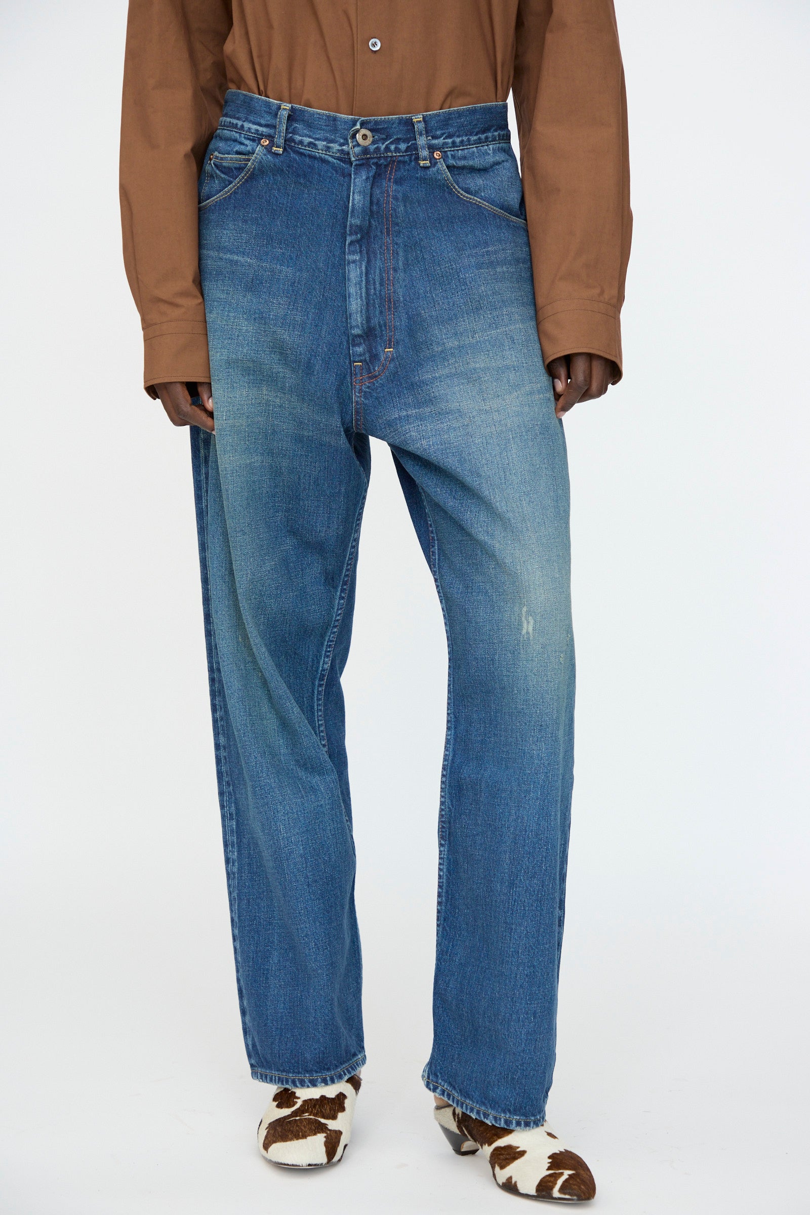 A person wearing a brown long-sleeve shirt, Chimala 13oz. Selvedge Denim Ranch Pants in Dark Wash, and cow-print pointed-toe shoes stands against a plain background.