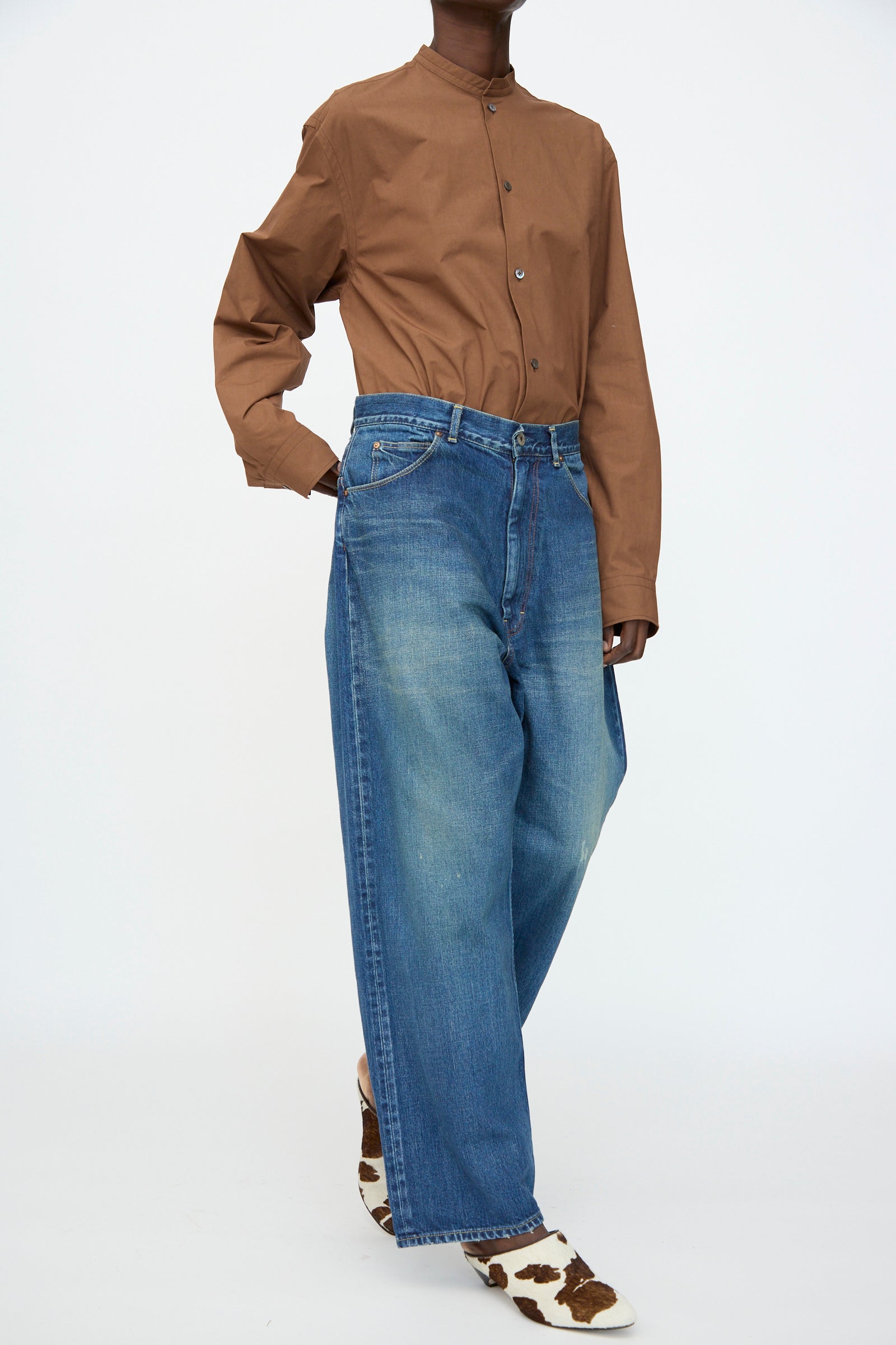 A person wearing a brown button-up shirt, Chimala 13oz. Selvedge Denim Ranch Pants in Dark Wash, and cow-print shoes stands against a plain white background.