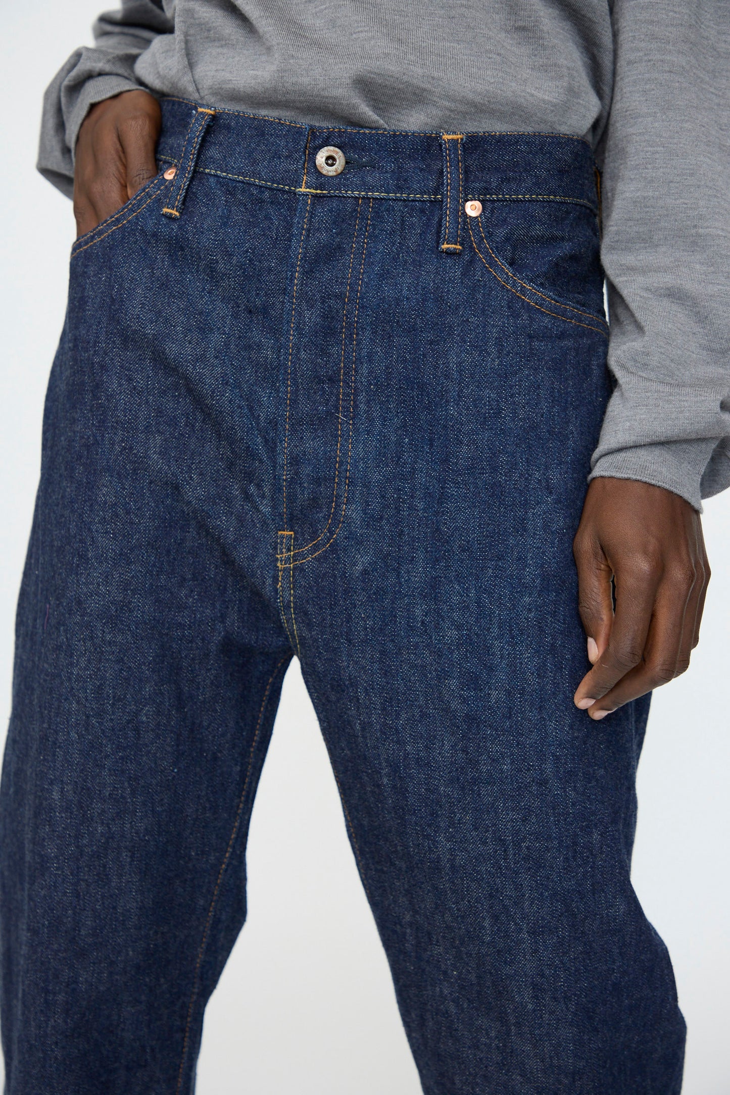 A person wearing a gray long-sleeve shirt and Chimala 13oz. Selvedge Denim Wide Tapered Cut in Rinse jeans, with one hand in their pocket and the other by their side.