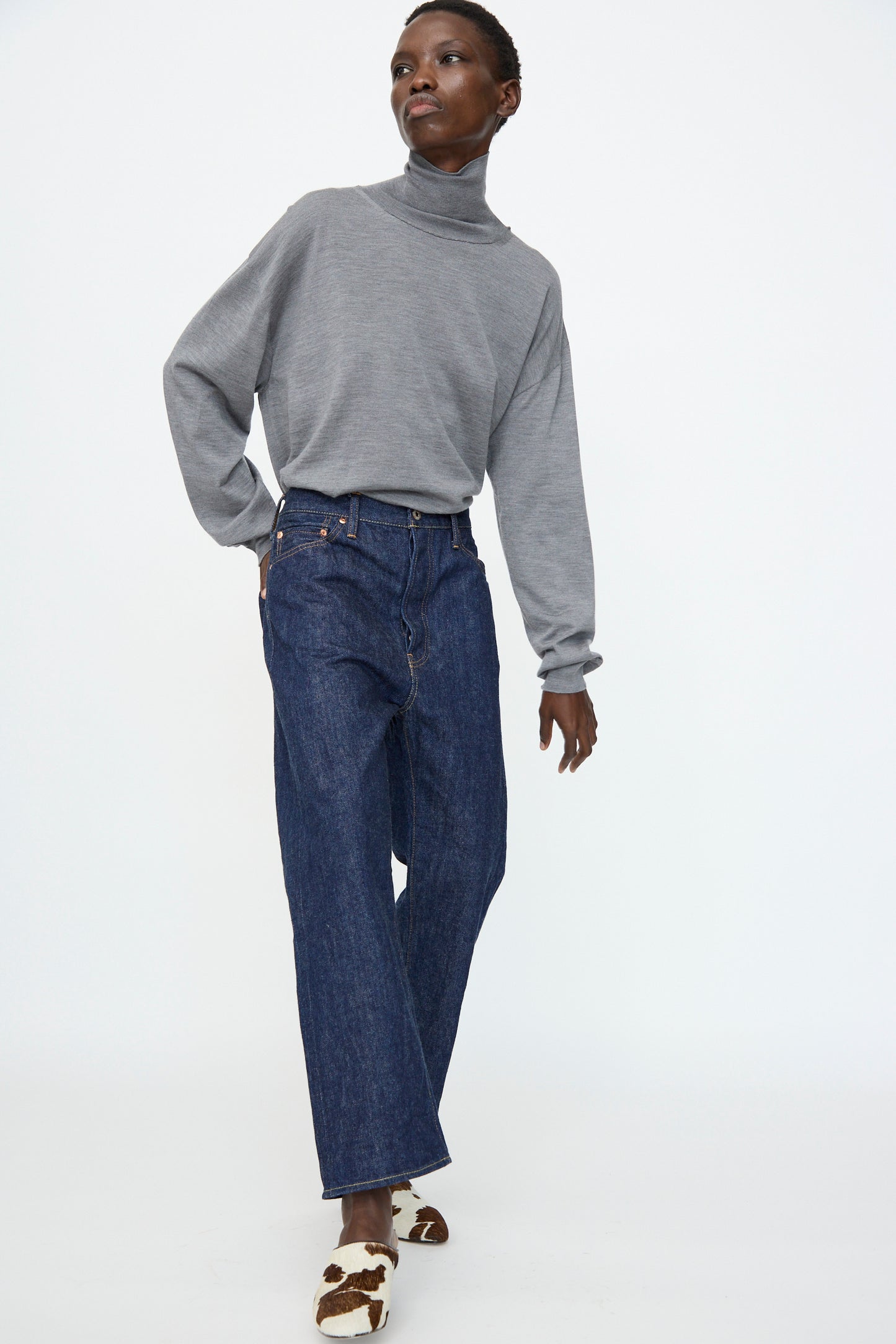 A person in a gray turtleneck sweater, Chimala 13oz. Selvedge Denim Wide Tapered Cut in Rinse jeans, and brown-and-white cow-print shoes stands against a plain white background.