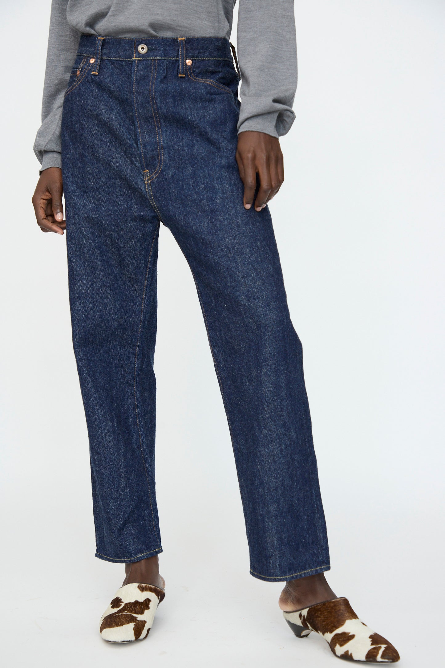 Wearing Chimala's 13oz. Selvedge Denim Wide Tapered Cut in Rinse jeans, a grey long-sleeve shirt, and brown-and-white cow print slip-ons, a person stands against a plain background.