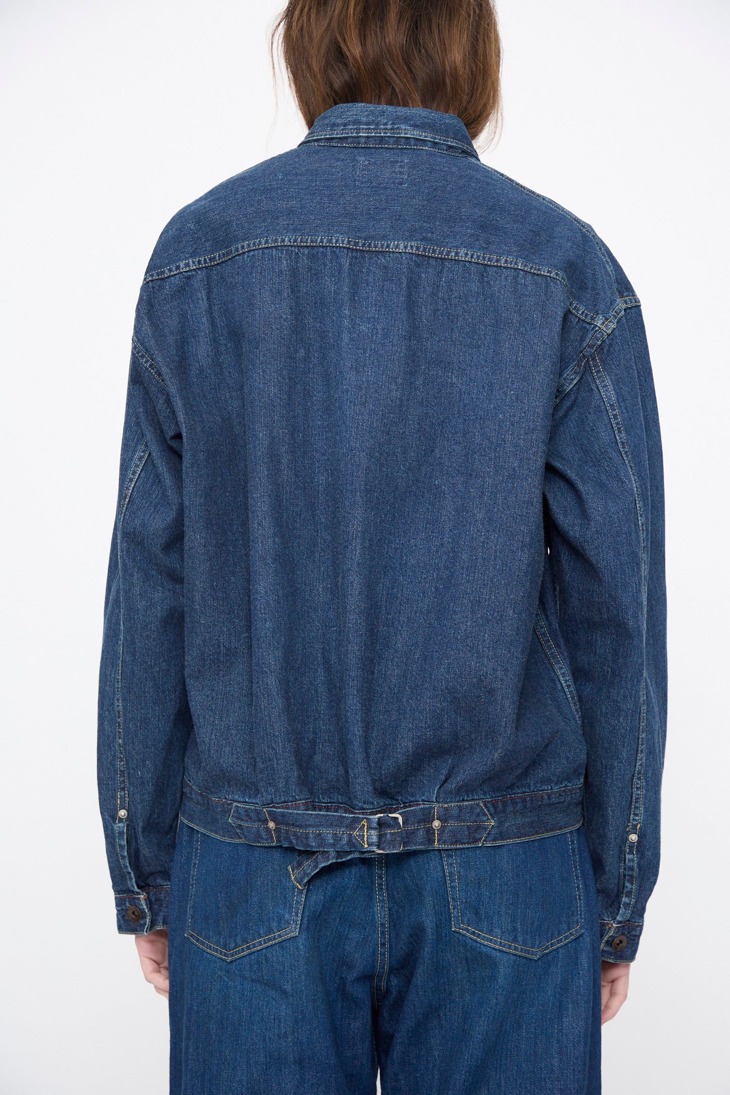 A person is shown from the back against a plain background, wearing Chimala’s Amoskeag Denim Truck Jacket in Dark Wash and blue jeans, both made in Japan.
