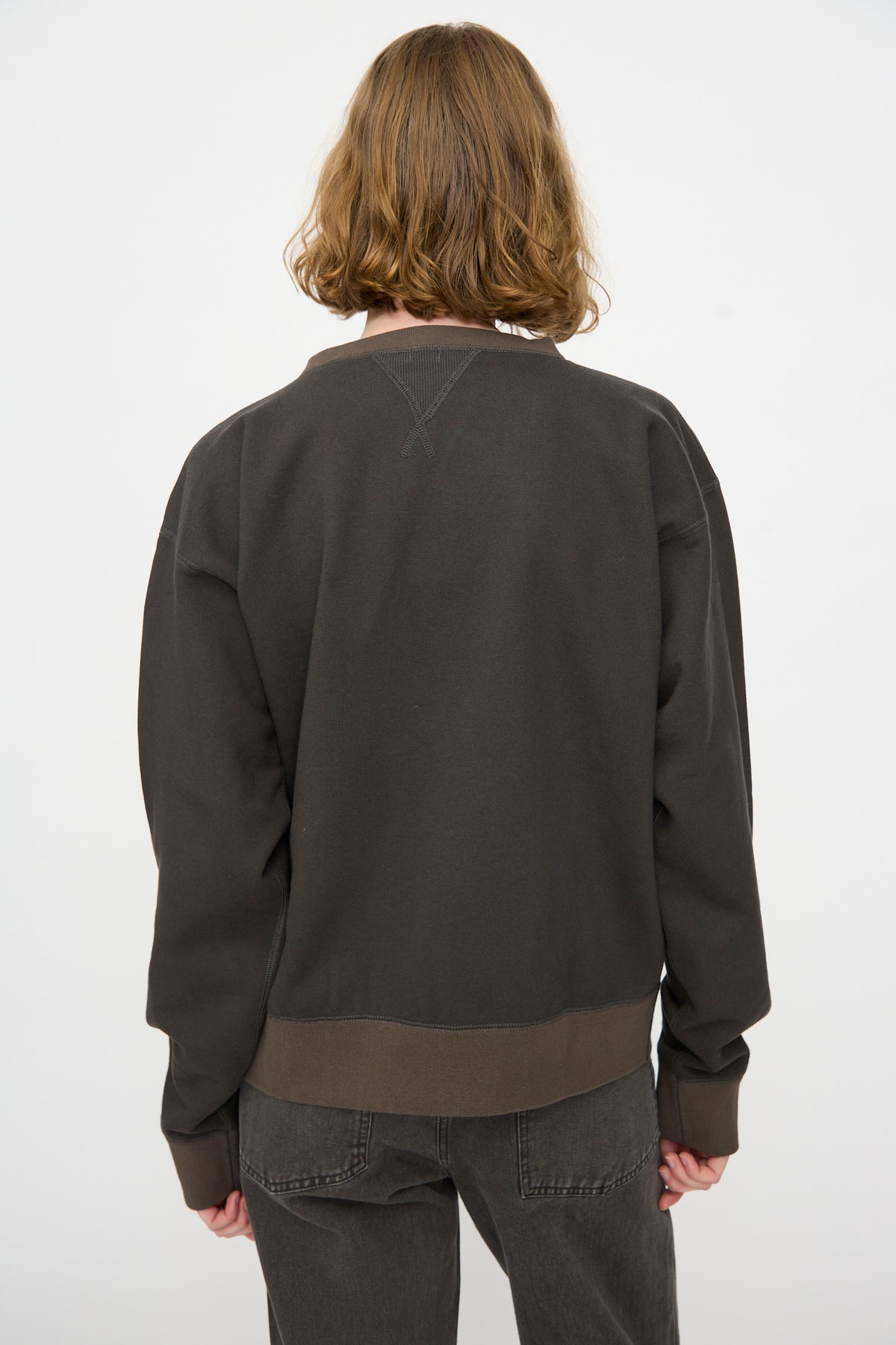 A person with shoulder-length brown hair is seen from the back, wearing the Chimala Brushed Fleece Crew Sweatshirt in Charcoal—a relaxed fit, long-sleeve top made from soft brushed cotton fleece—paired with dark jeans and standing against a plain white background.