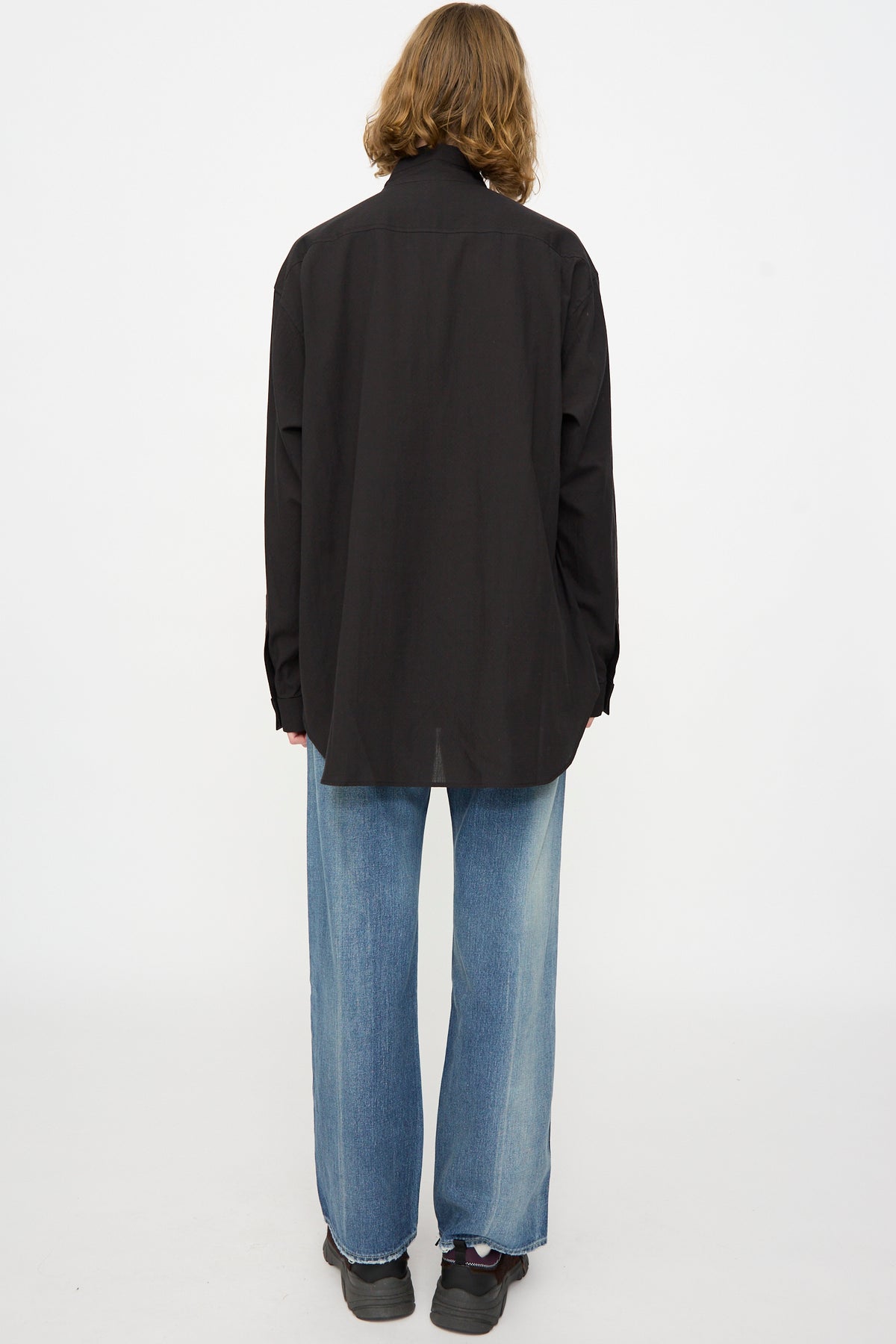 A person with shoulder-length hair, seen from behind, wears the Chimala Cotton Dobby Bow Tie Blouse in Black, paired with light blue wide-leg jeans and black shoes, standing against a plain white background.
