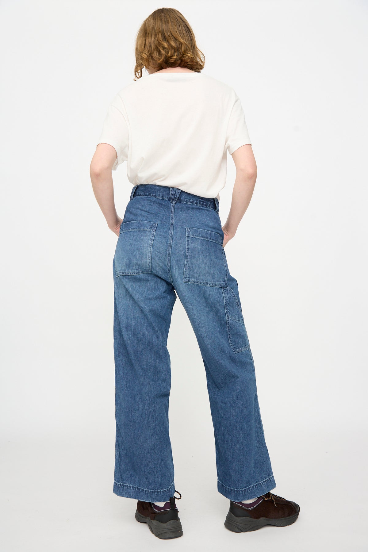 A person stands with their back to the camera, wearing a white shirt, Chimala Denim Painter Pant in Medium Engineer featuring a relaxed wide-leg fit, and dark shoes, set against a plain white background.
