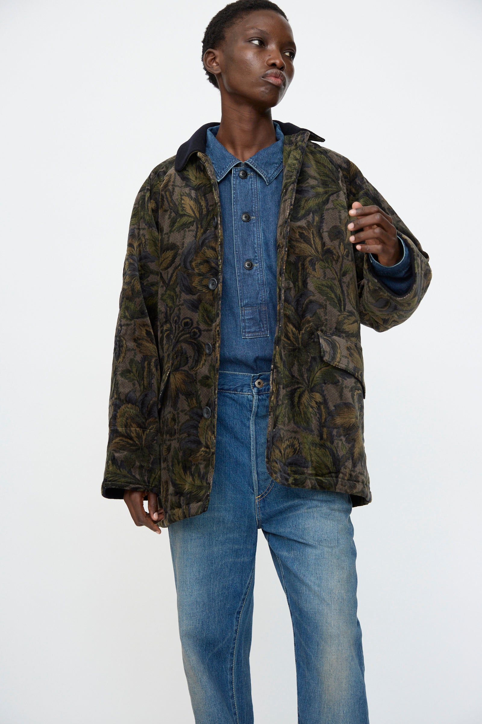 Dressed in Chimala's Floral Cord Barn Coat in Black over a denim shirt and blue jeans, the person stands confidently against a plain white background.