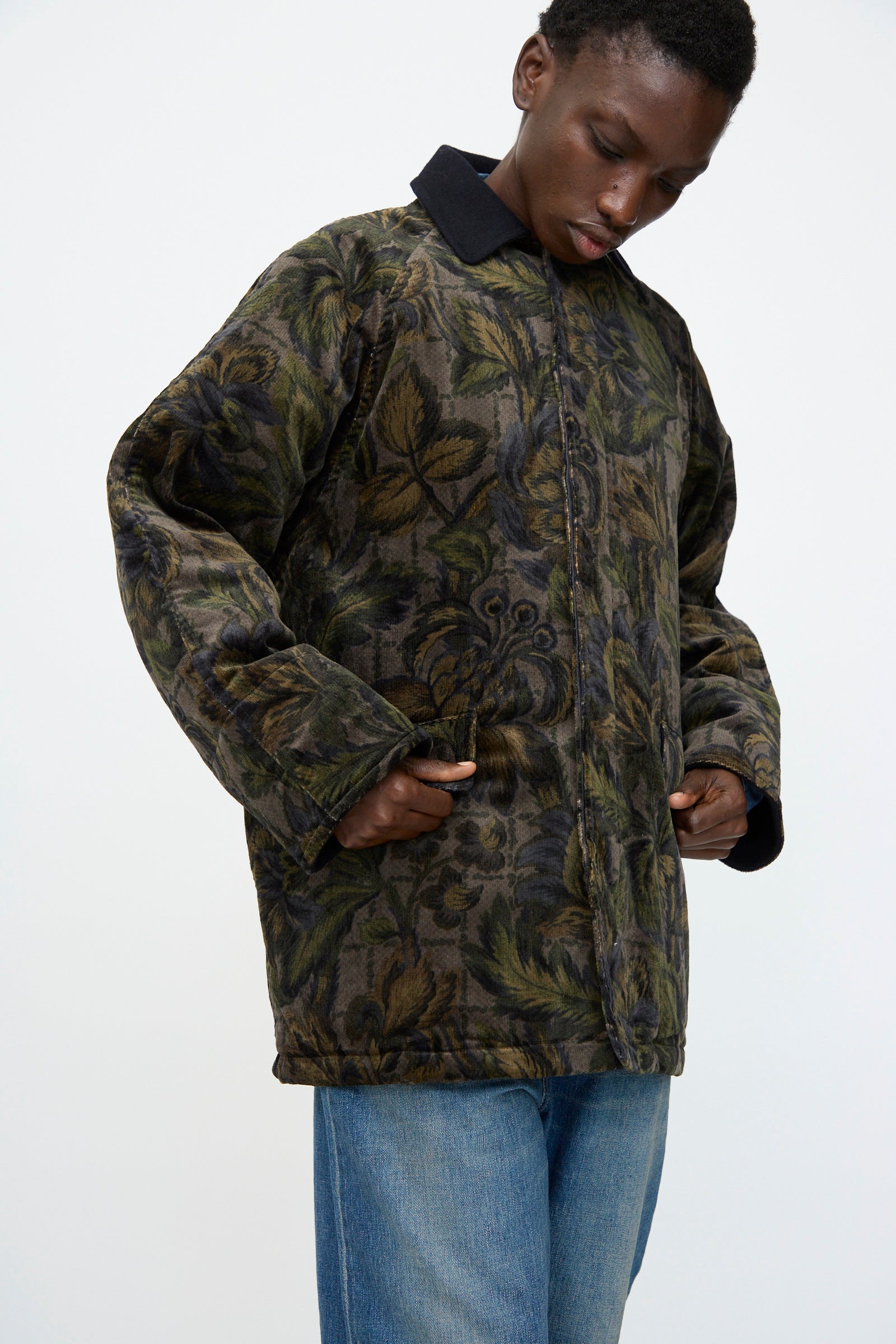 A person wearing the Chimala Floral Cord Barn Coat in Black with blue jeans, hands in pockets, looks down against a plain background.