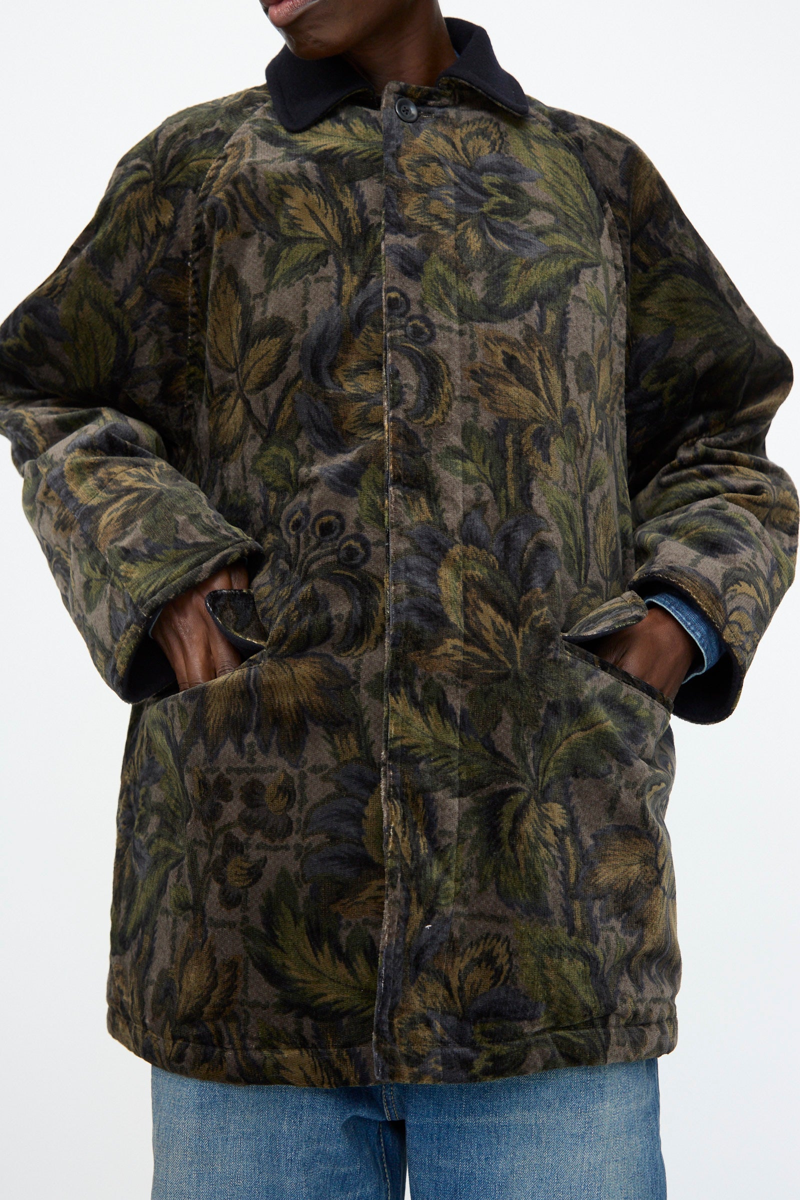 A person wearing the Chimala Floral Cord Barn Coat in Black with front pockets, blue jeans, and a black collar stands with hands in pockets against a plain background.