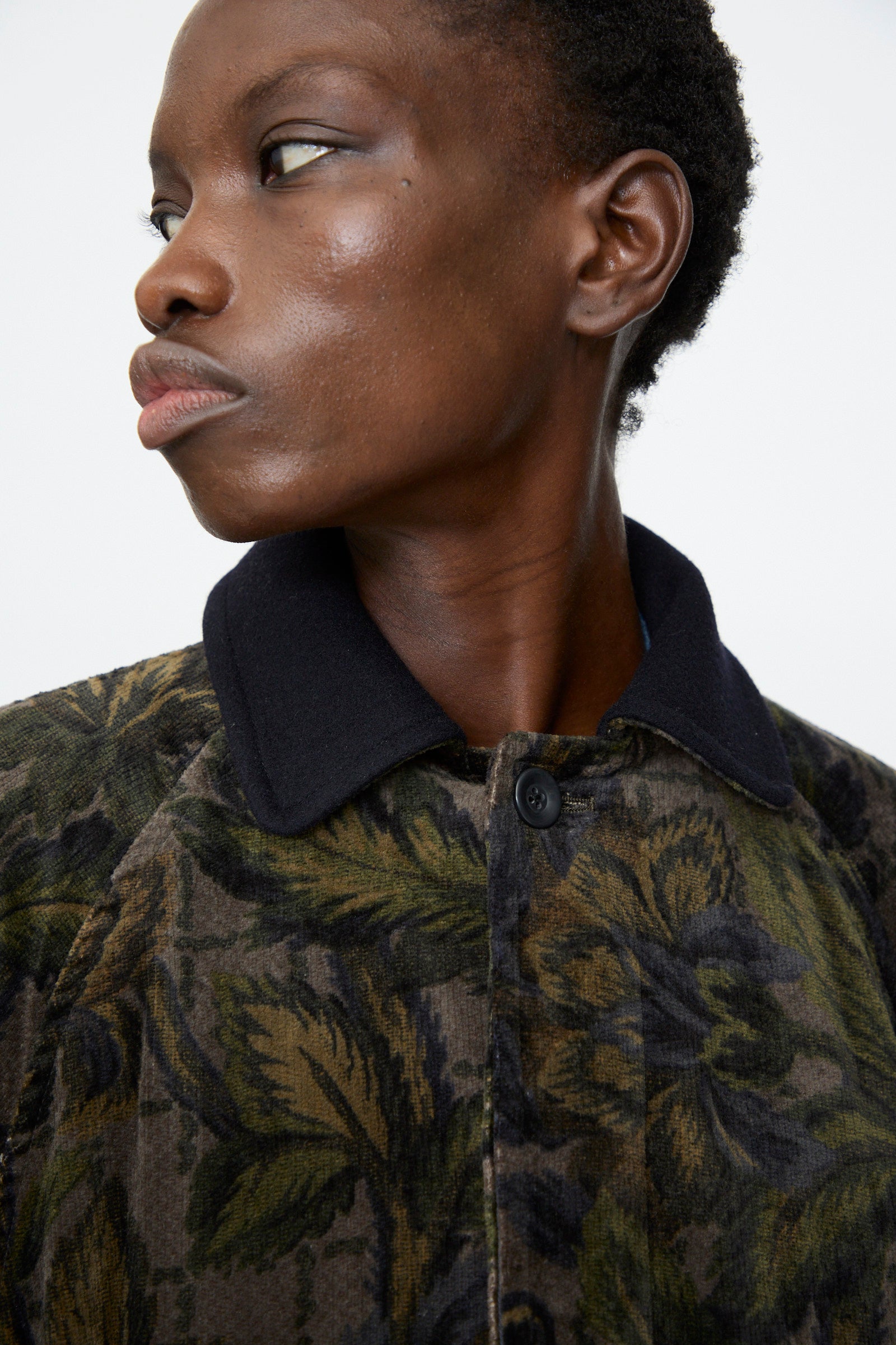 A person wears the Chimala Floral Cord Barn Coat in Black, featuring a patterned design and black collar, while looking to the side against a plain background.