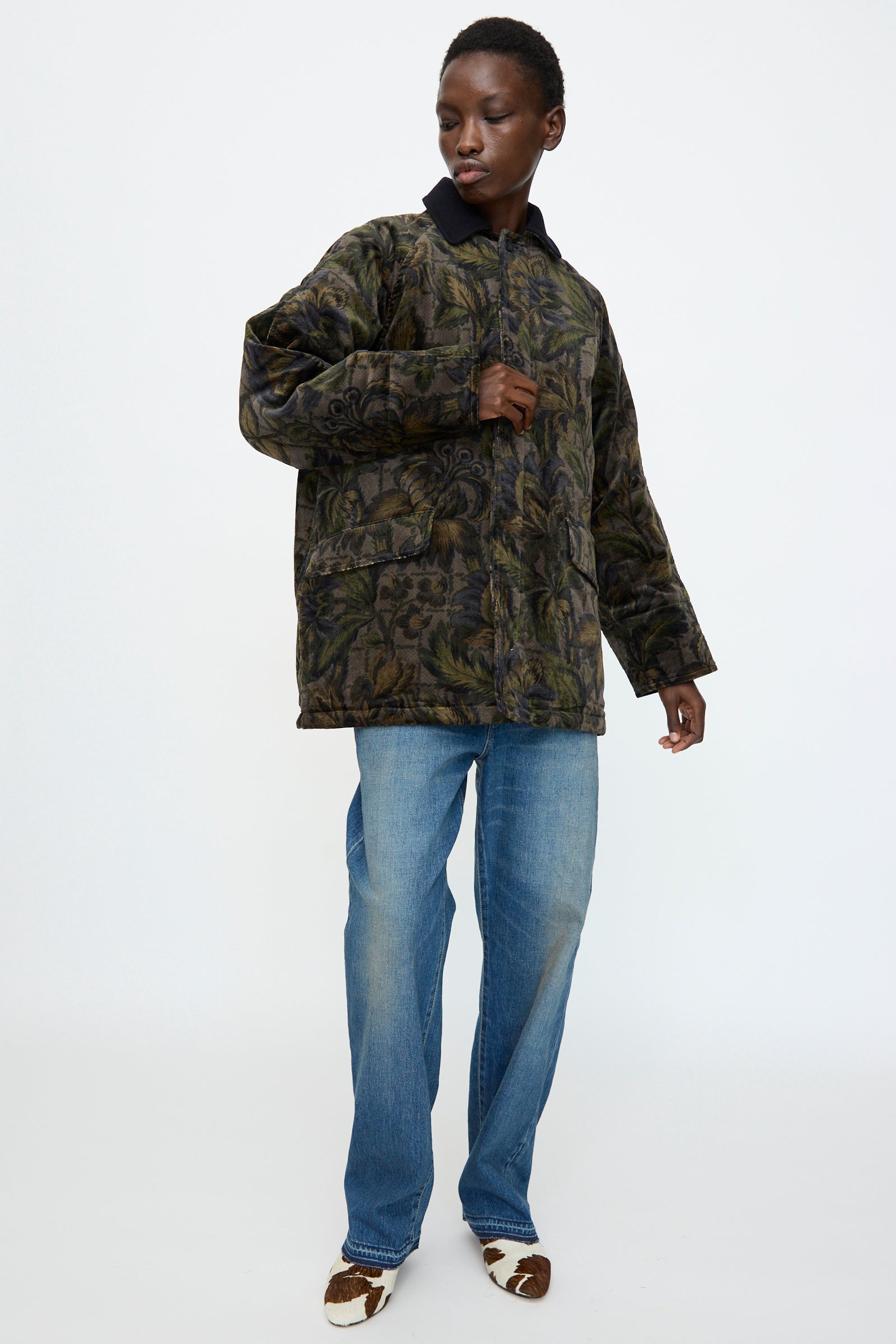 A person stands against a plain background wearing the Chimala Floral Cord Barn Coat in Black, paired with blue jeans and brown-and-white shoes for a relaxed yet distinctive look.