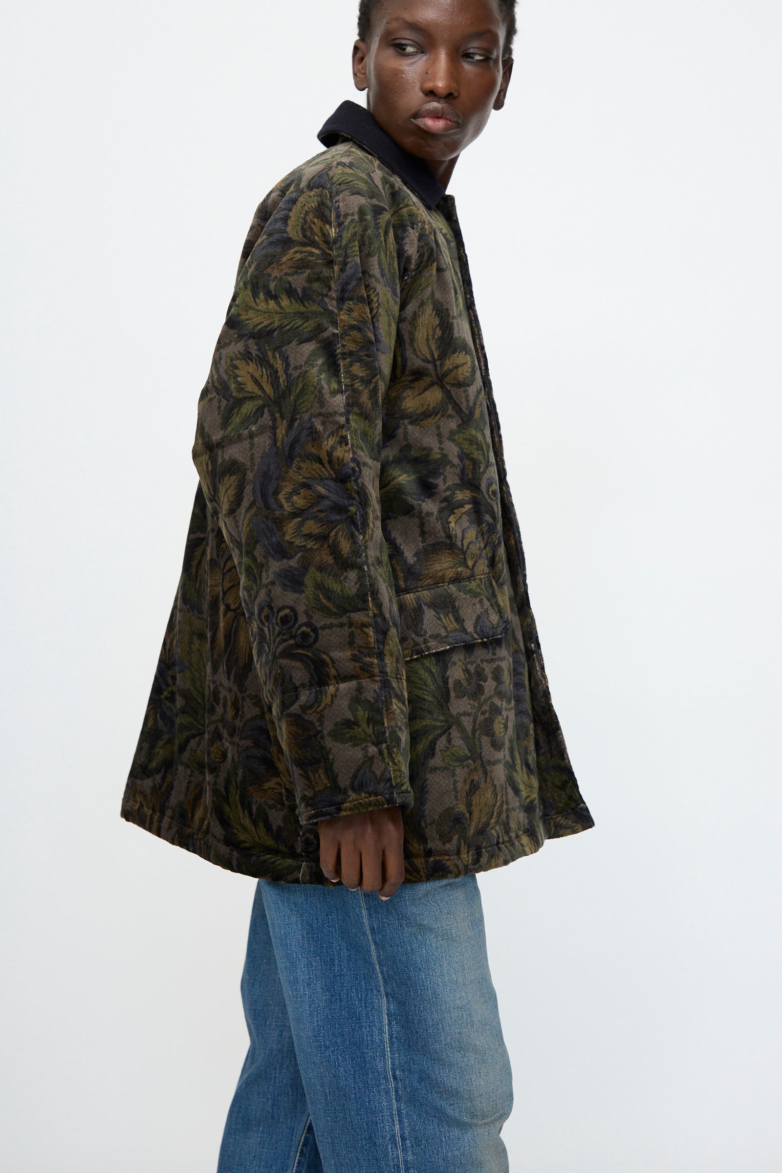 A person wears the Chimala Floral Cord Barn Coat in Black with leaf designs and blue jeans, glancing over their shoulder against a plain light background.