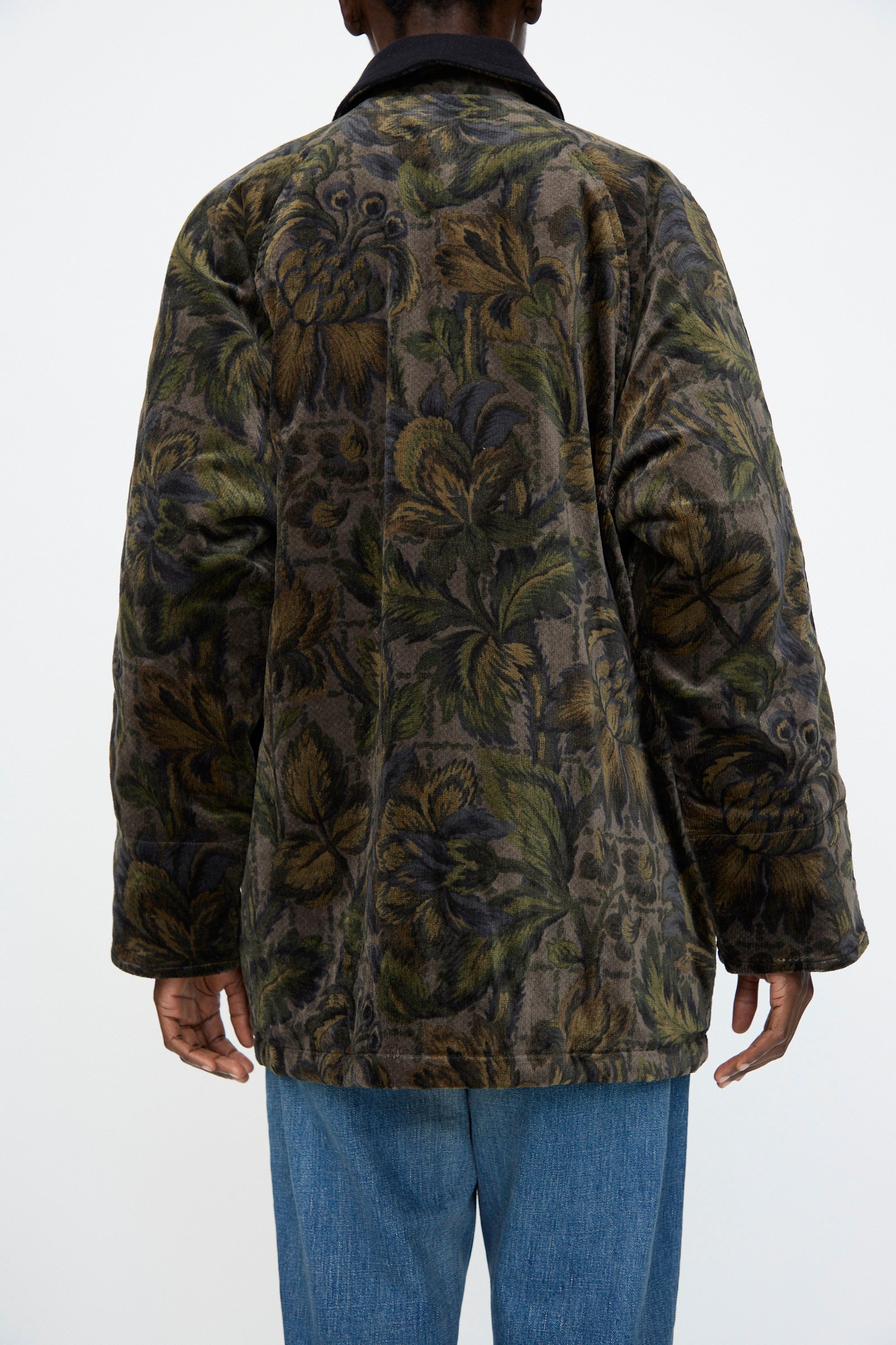 A person is seen from behind against a light background, wearing Chimala’s Floral Cord Barn Coat in Black paired with blue jeans.
