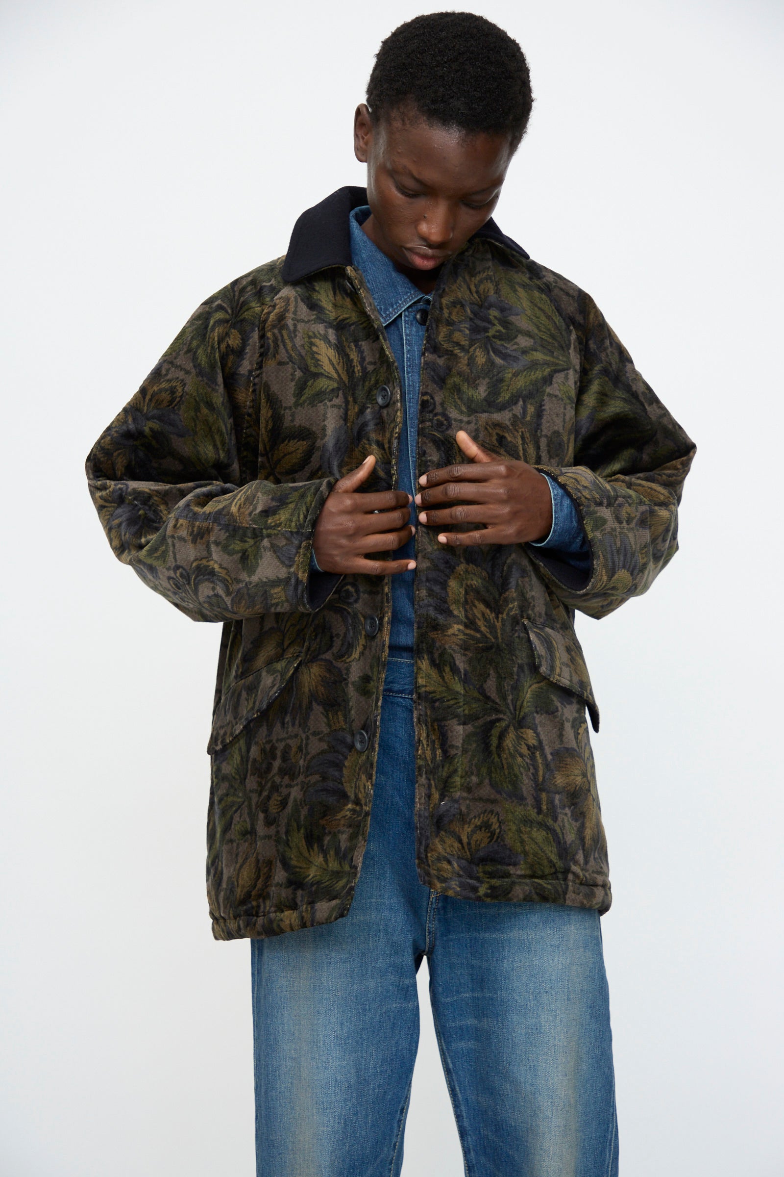 A person in the Chimala Floral Cord Barn Coat in Black—crafted from floral corduroy and denim, made in Japan—stands looking down and adjusts the front of the jacket.