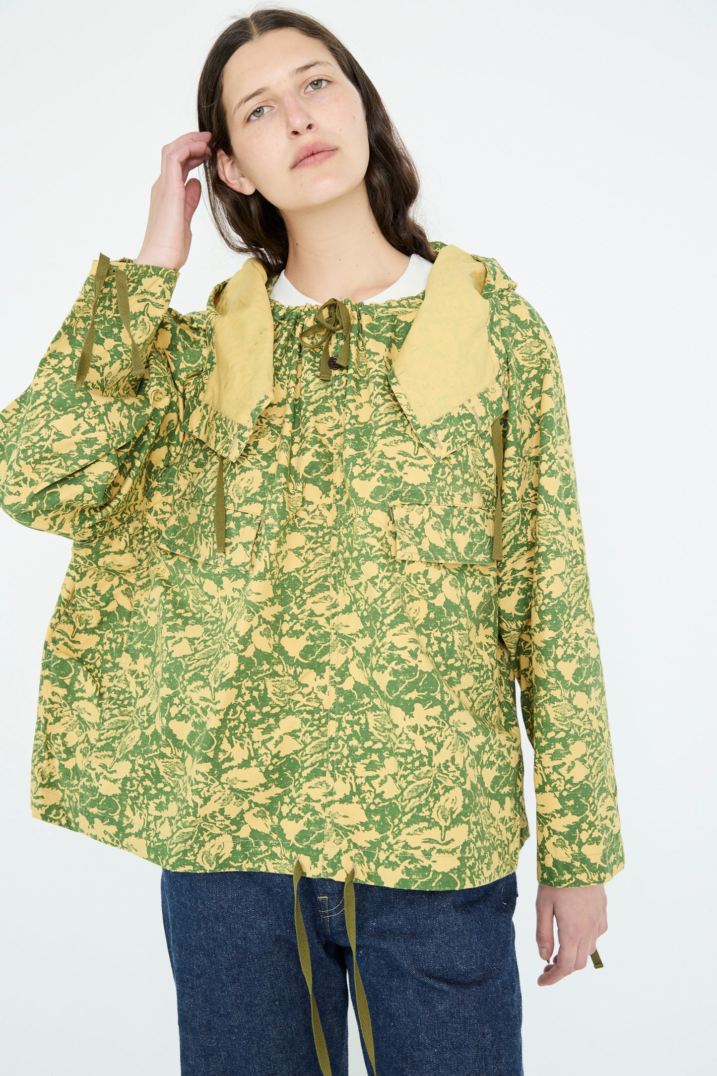 Against a plain background, a person with long dark hair gently touching their head is wearing Chimala's Summer Leaf Smock Jacket in Camo, which features a green and beige floral pattern, along with jeans.