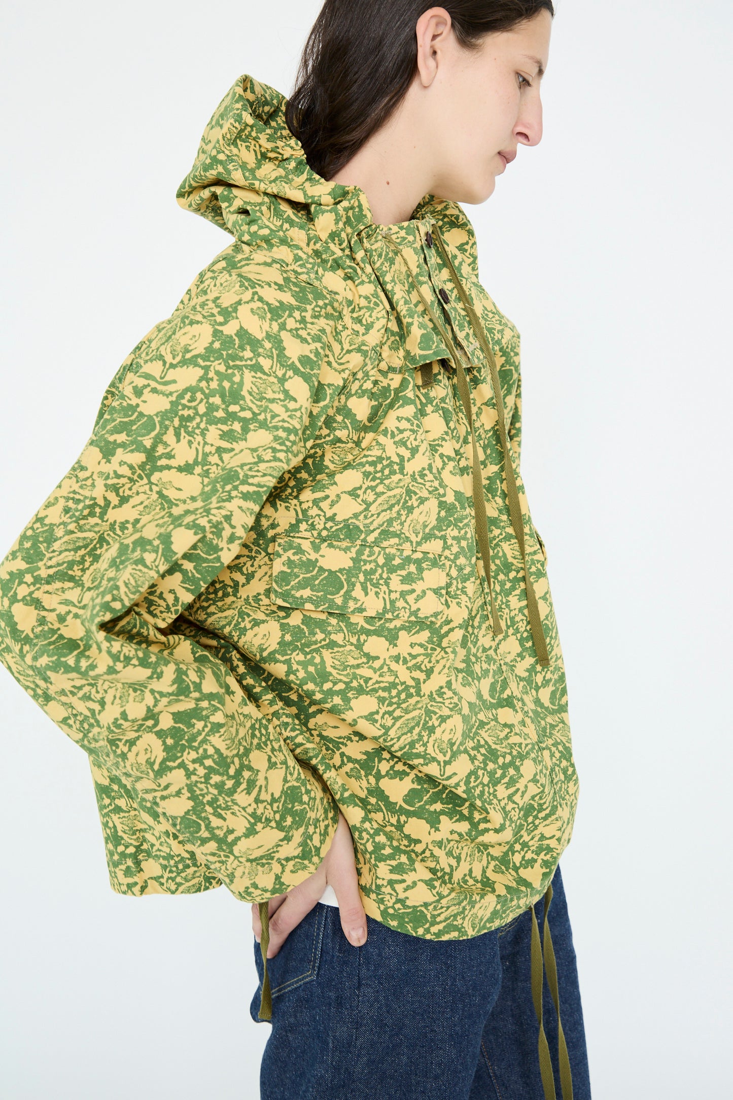 A person stands with hands in pockets, wearing the Chimala Summer Leaf Smock Jacket in a green and yellow camo pattern, paired with jeans against a plain background.